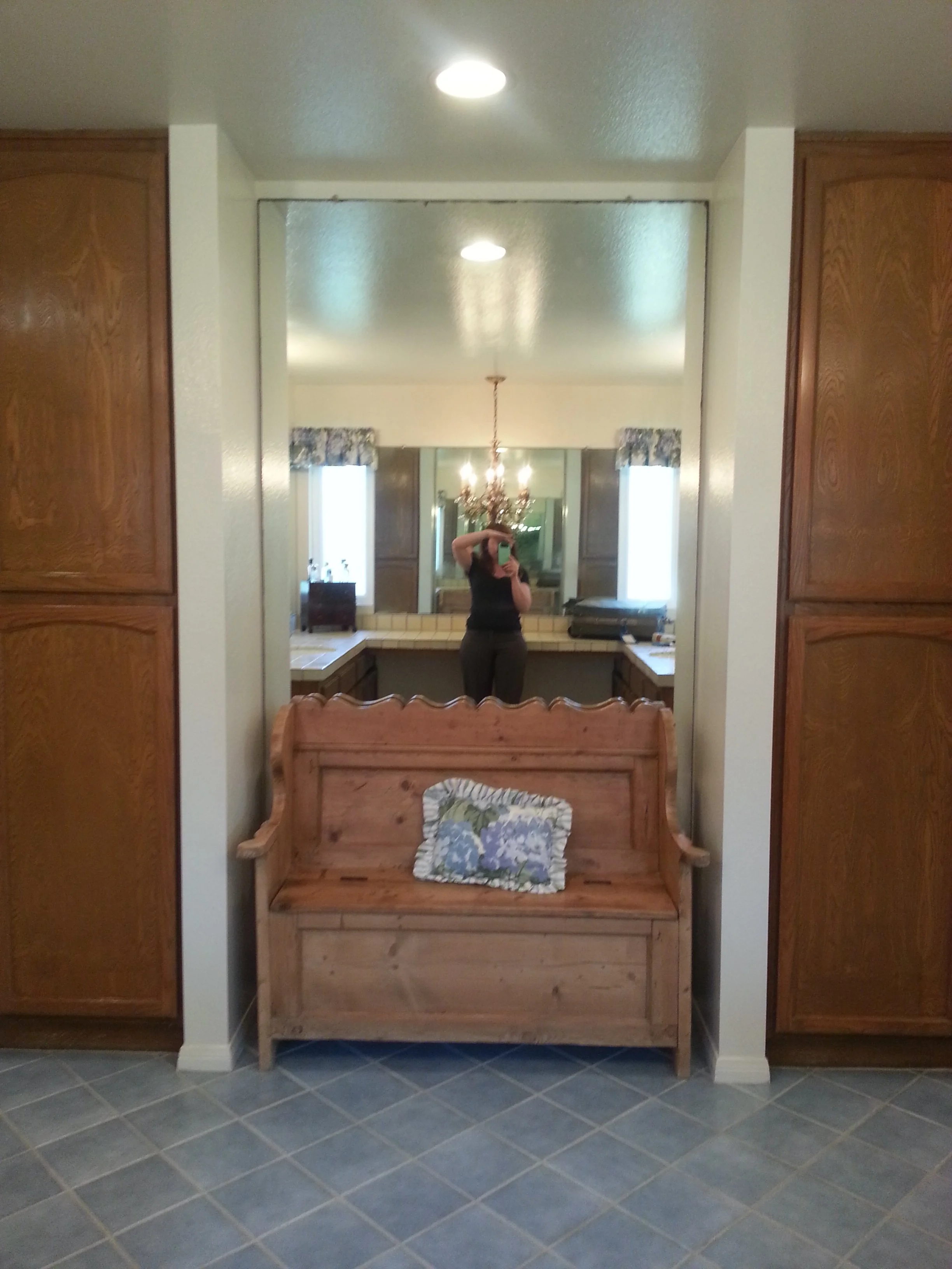 A large rounded mirror above a wooden bench with a floral pillow, flanked by wooden cabinets. A person is visible taking the photo, reflected in the mirror, with a chandelier hanging from the ceiling and two windows with blue curtains in the backgrou