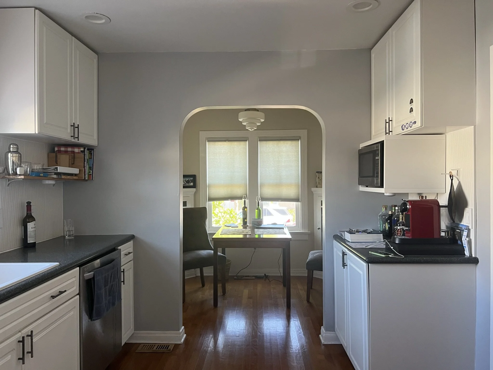 A kitchen with white cabinets, black countertops, and a dining area near two windows with blinds, natural light, and a wooden floor.