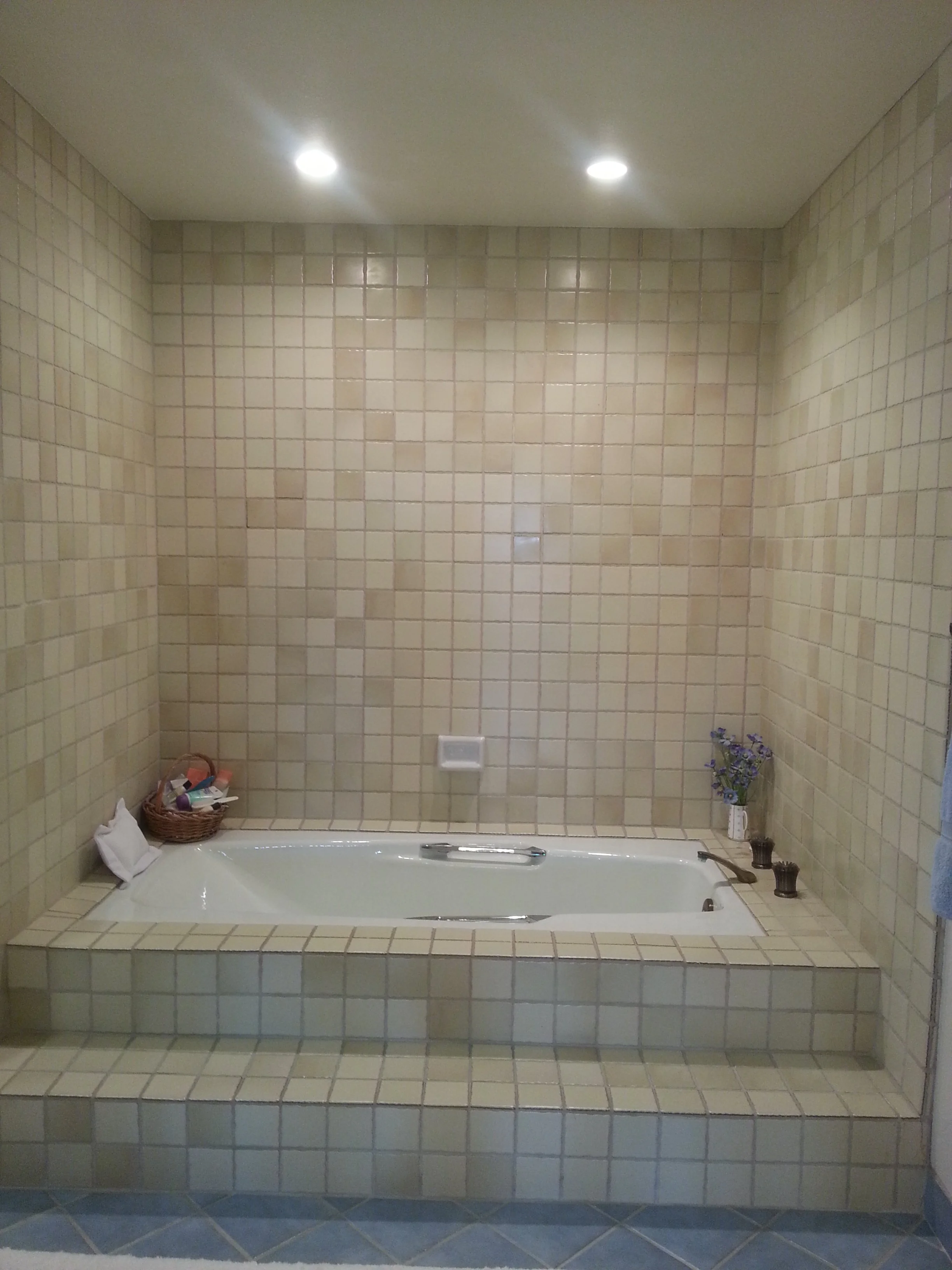 A built-in bathtub with beige tiled surround in a bathroom. The tiles extend to the ceiling behind the tub. There is a basket with toiletries and a white towel on the edge of the tub, and a small vase with purple flowers on the right side.