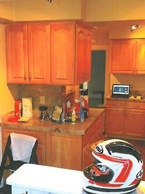 A kitchen with wooden cabinets, a countertop with various items including bottles and containers, a black chair with a white cloth draped over it, a white table, and a motorcycle helmet on the table.