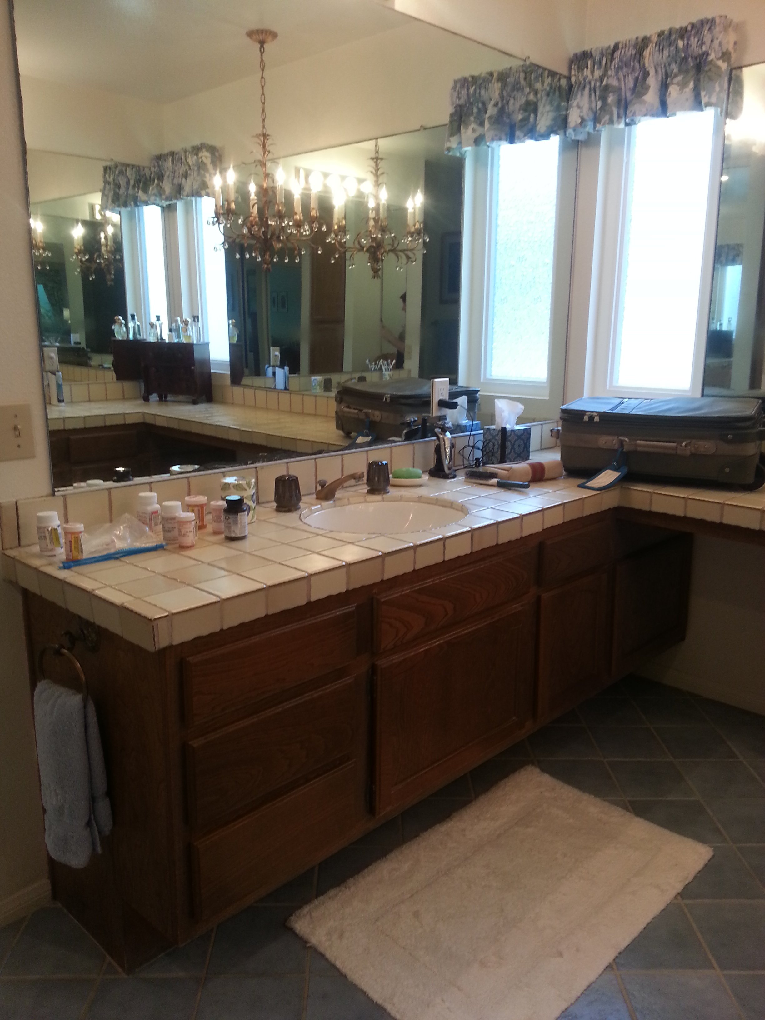 A bathroom vanity with a tiled countertop, medicine bottles, a toothbrush holder, and a tissue box. There is a large mirror above the vanity, a chandelier hanging from the ceiling, and two windows with floral valances. A beige bathmat is on the tiled
