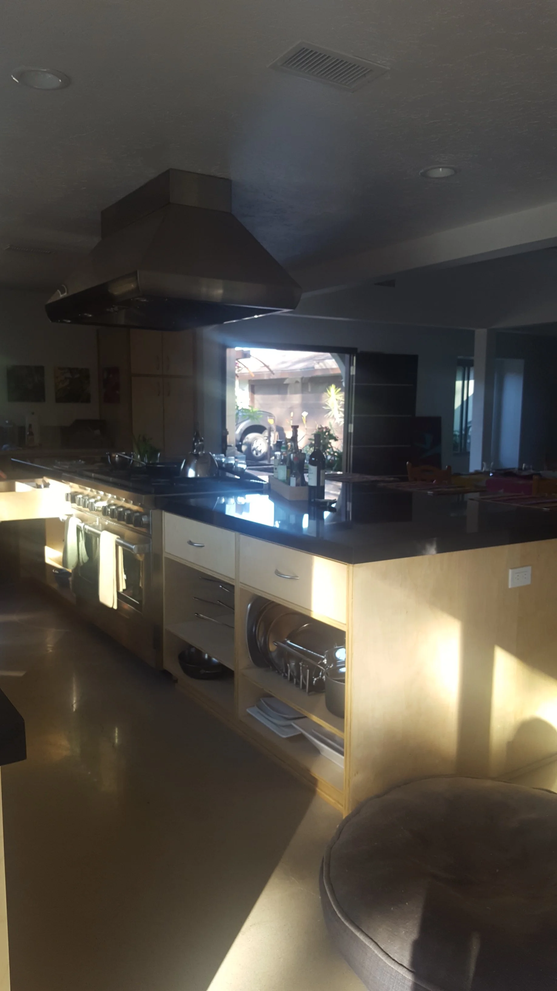 A modern kitchen with a black range hood, wooden cabinets, a black countertop, and a window letting in natural light. There are bottles and kitchenware on the counter, and a gray upholstered round chair in the foreground.