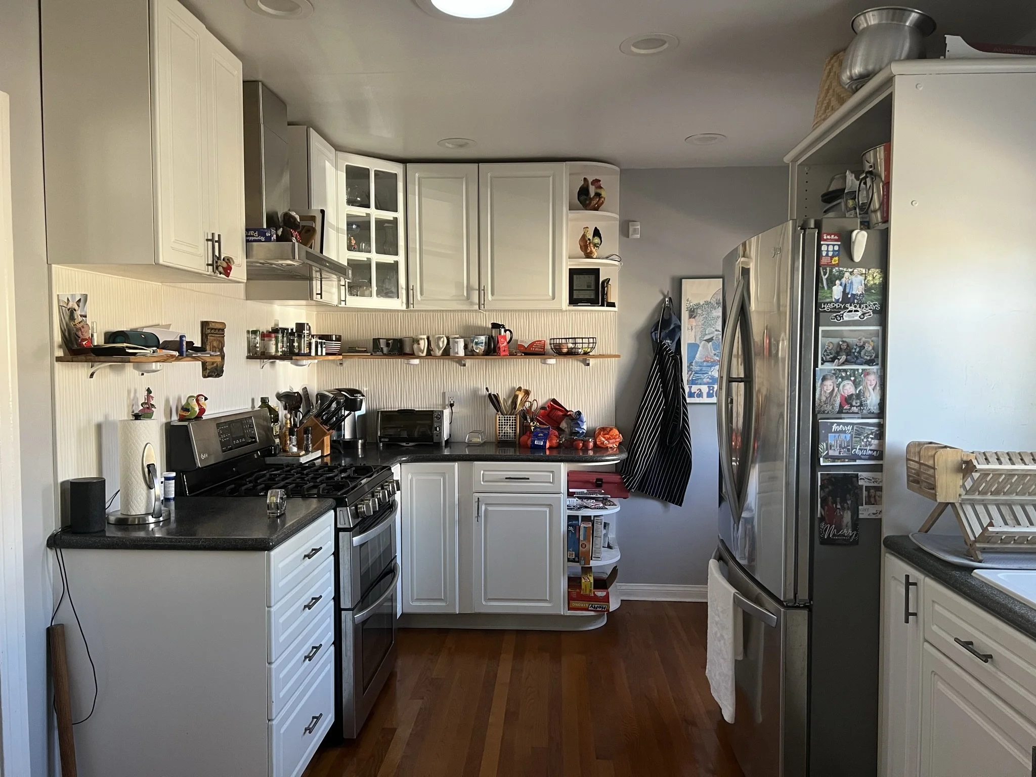 A kitchen with white cabinets, black counters, stainless steel appliances, and various items on the counter including coffee mugs, a toaster oven, and a microwave. There are decorative rooster figures on a shelf and framed pictures on the wall.