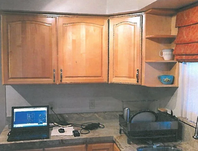 Kitchen countertop with a laptop, various electronics, and a drying rack, wooden upper cabinets, and a window with an orange shade.