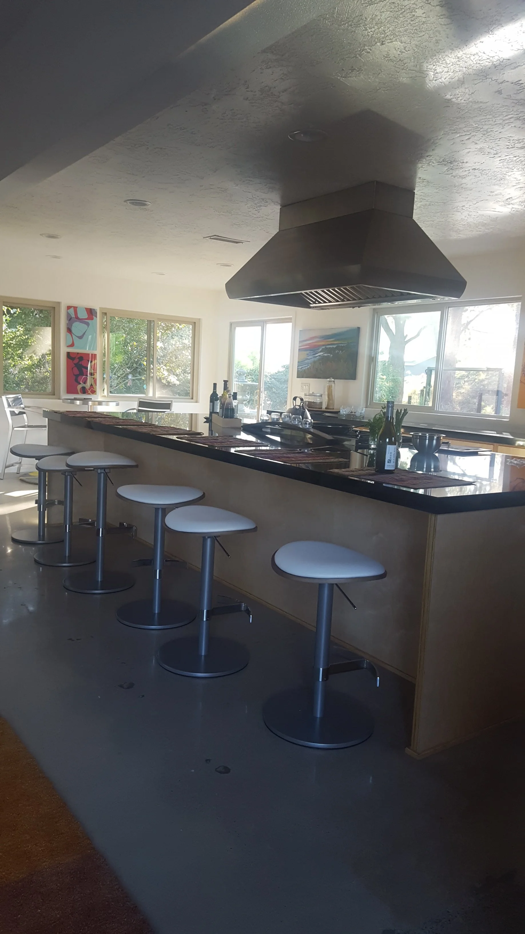 A modern kitchen with a black countertop bar and white bar stools, large windows, and contemporary artwork on the wall.