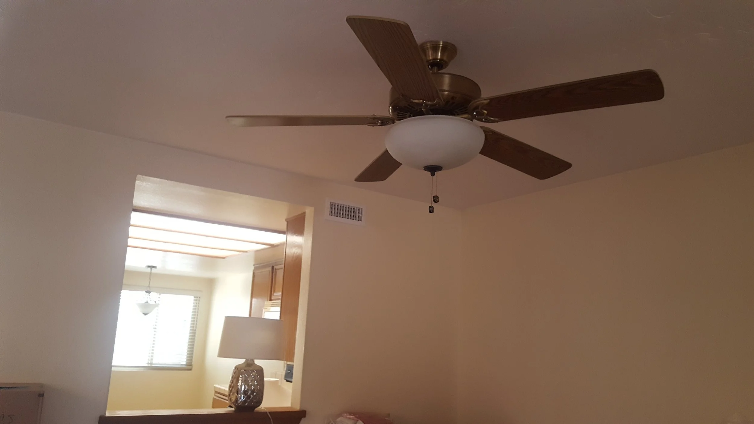 Living room with a ceiling fan and a light fixture, kitchen is visible through an opening with wooden cabinets and window blinds.