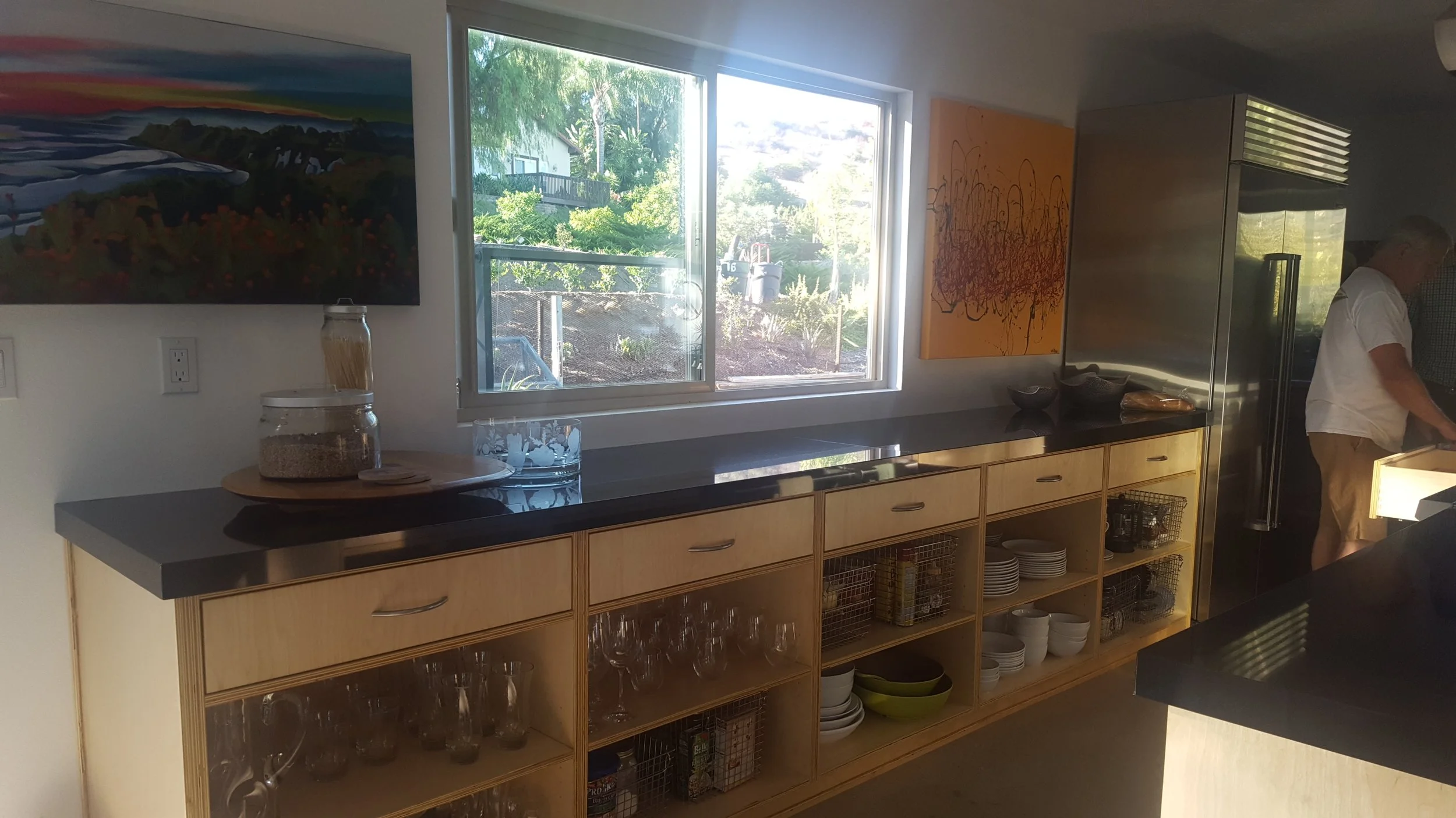 Kitchen area with wooden cabinets, black countertop, and open shelves containing glassware, plates, and bowls. A window showing trees and part of a garden. Artworks on the wall and a person in white shirt and khaki shorts near the fridge.