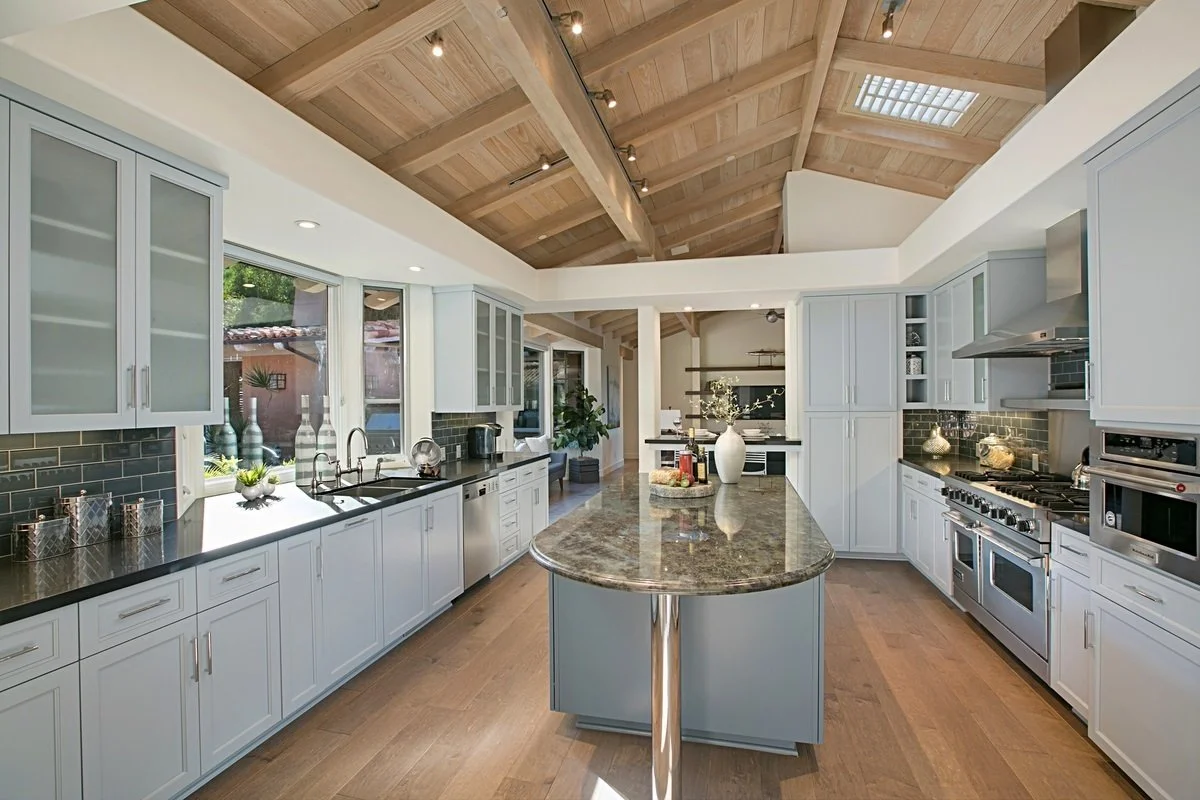 Modern kitchen with white cabinets, black countertops, a central island with a granite top, and stainless steel appliances. Large windows let in natural light, and a vaulted wooden ceiling adds warmth to the space.