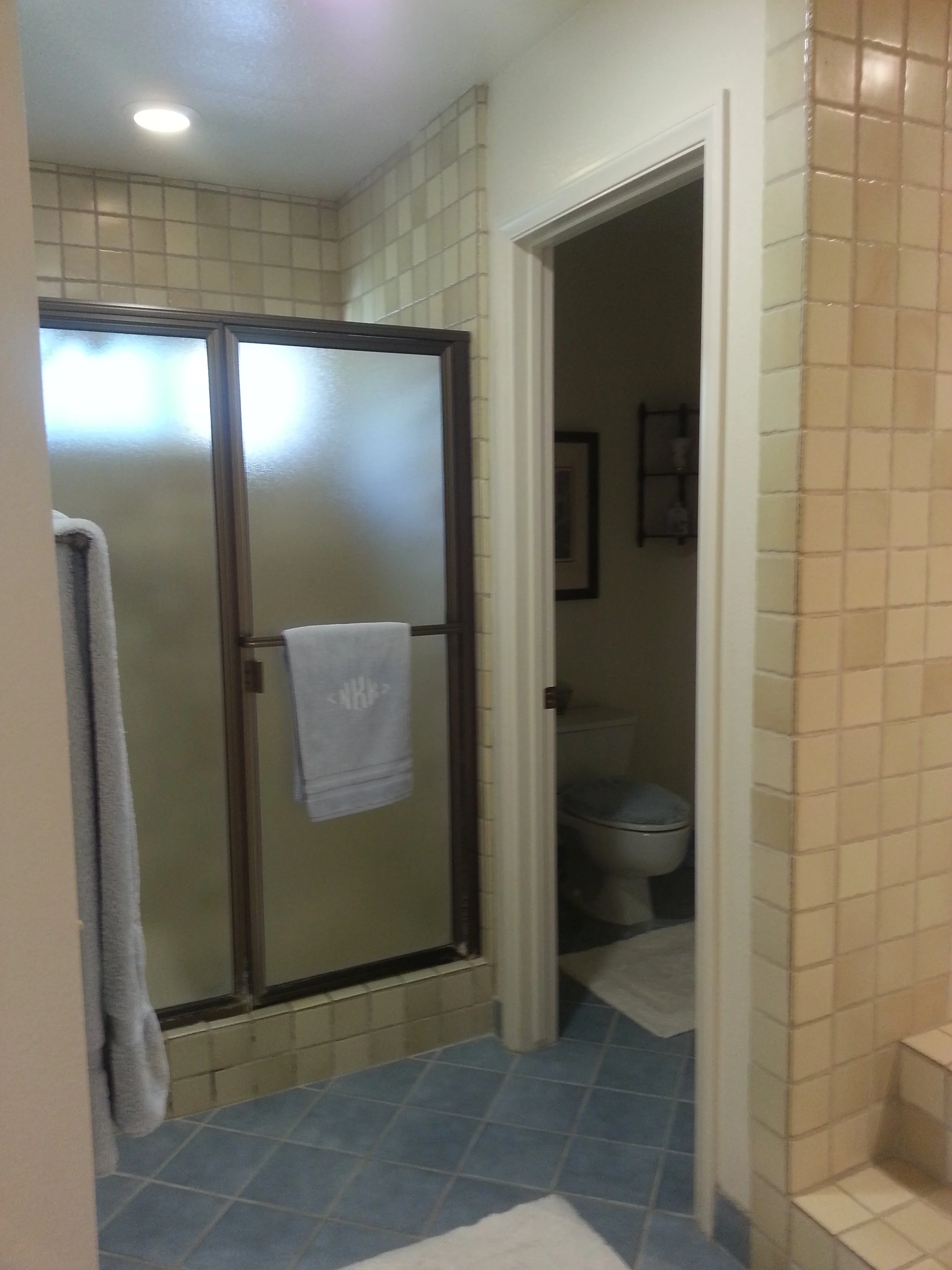 Bathroom with a shower stall with frosted glass door, a towel hanging on the door, a toilet visible to the right, beige tiled walls, and blue tiled floor.