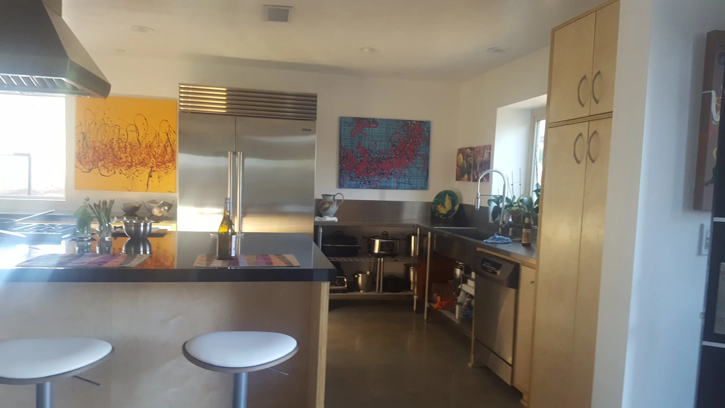 Modern kitchen with light wood cabinets, stainless steel appliances, colorful abstract paintings on the wall, a central island with a black countertop, and two white bar stools.