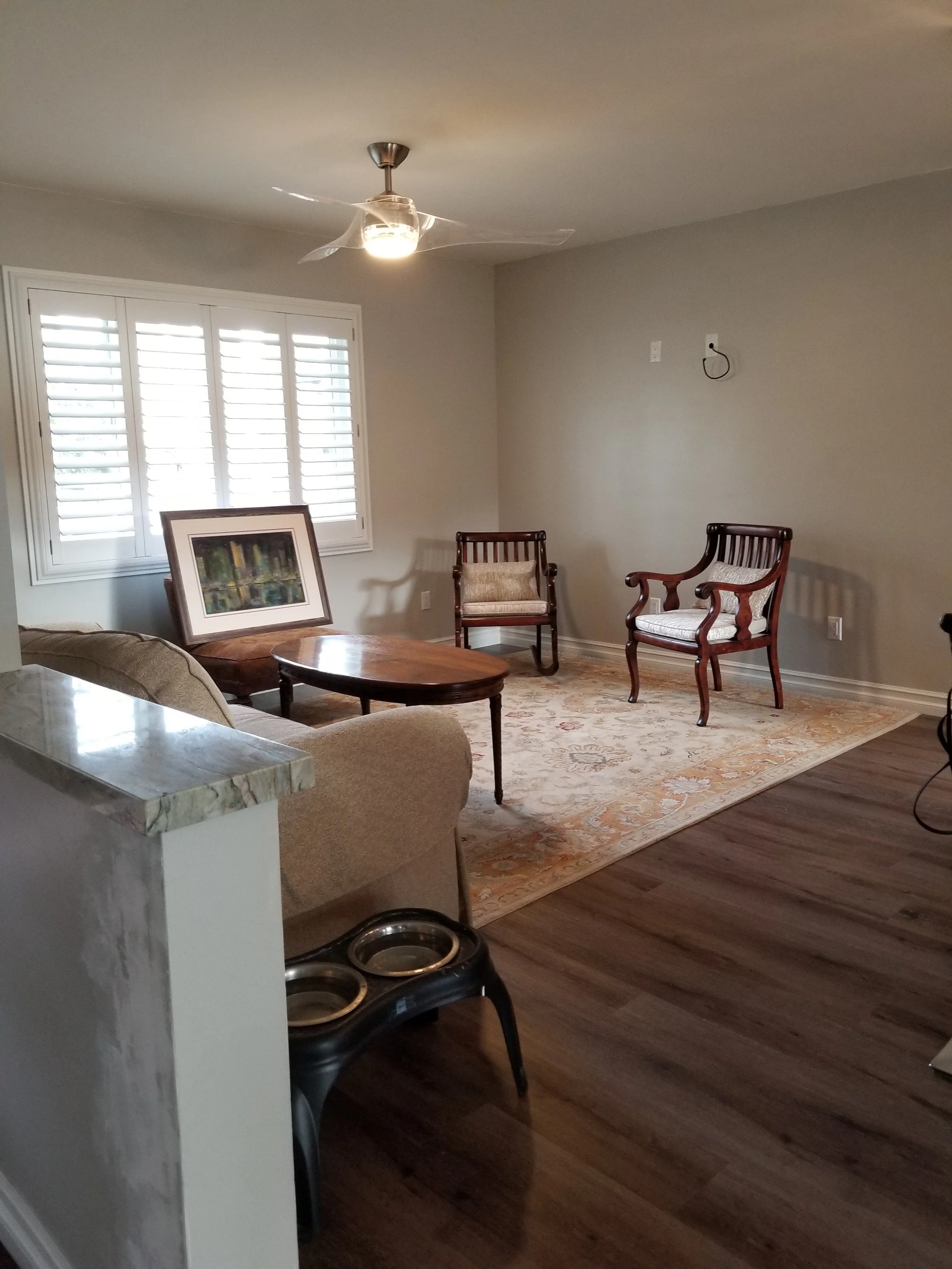 Living room with a ceiling fan, window with shutters, wooden chairs, a framed artwork, and a patterned area rug.