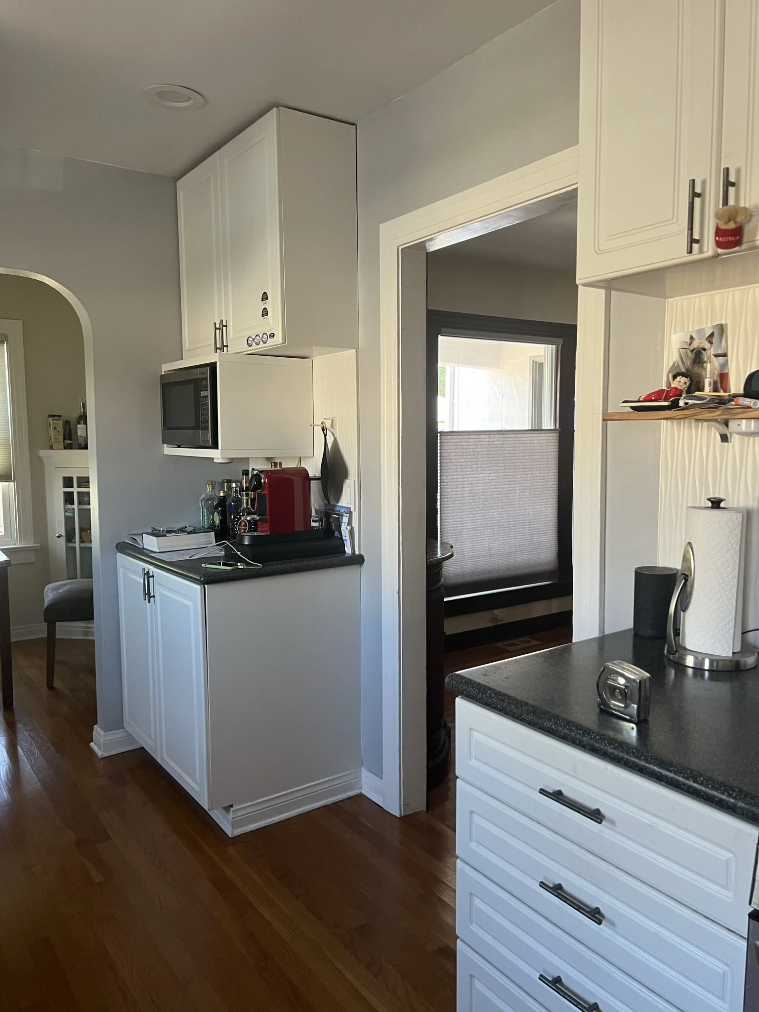 Kitchen with white cabinets, a stove, a microwave, and a small bathroom with a towel and other items on a shelf. There are wooden floors and a doorway leading to another room with a window.