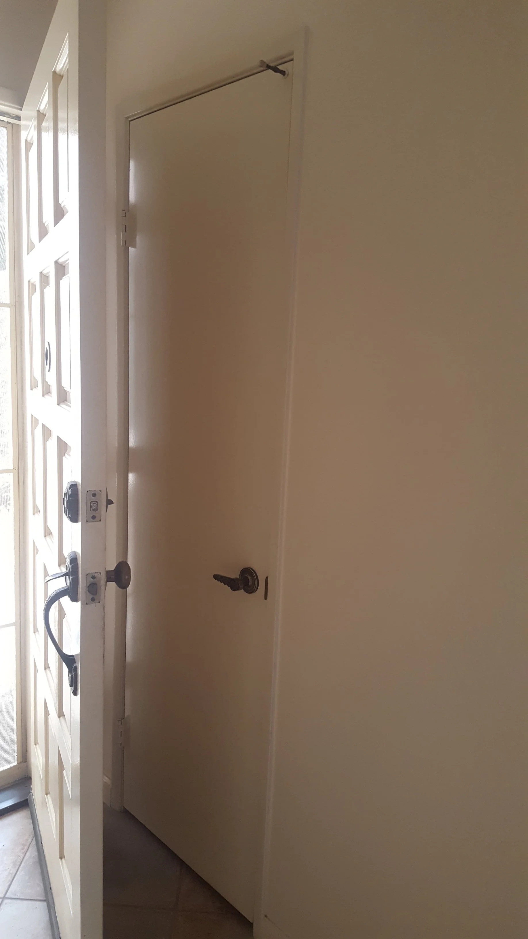The image shows a white door with a security screen in front of it, with a deadbolt lock, handle, and a key in the lock. The door is slightly open, revealing part of the porch or hallway outside.