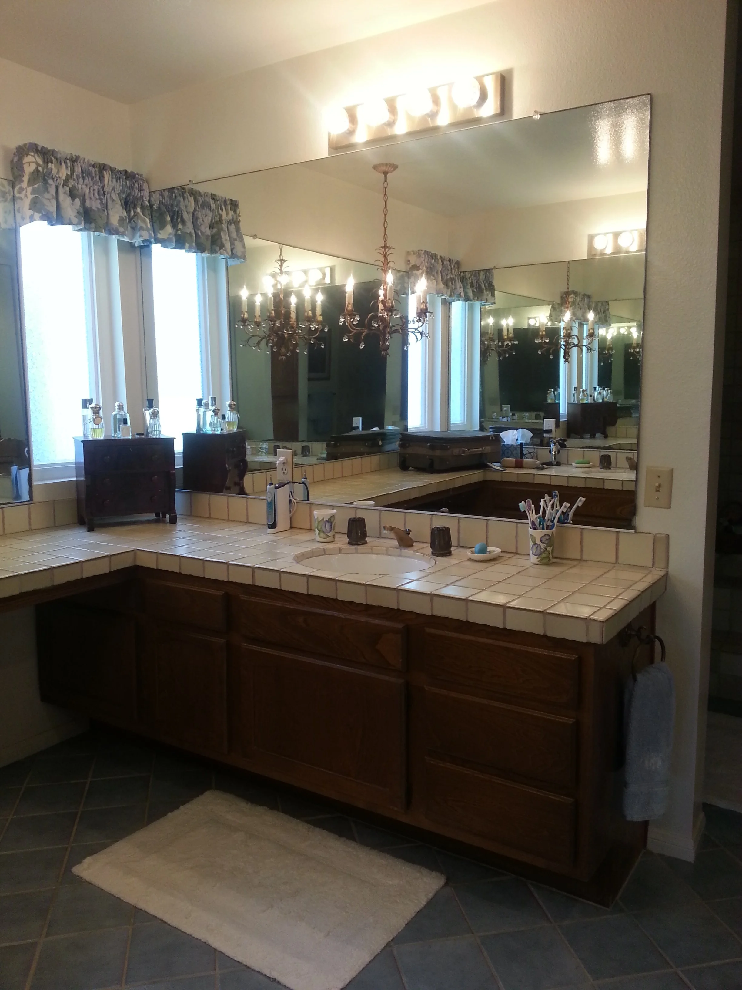 Bathroom vanity with a large mirror, a chandelier light fixture, a sink, and various toiletries. There are two small dark wooden cabinets and a cream-colored rug on the gray tiled floor. The window has floral curtains and multiple bottles on the wind