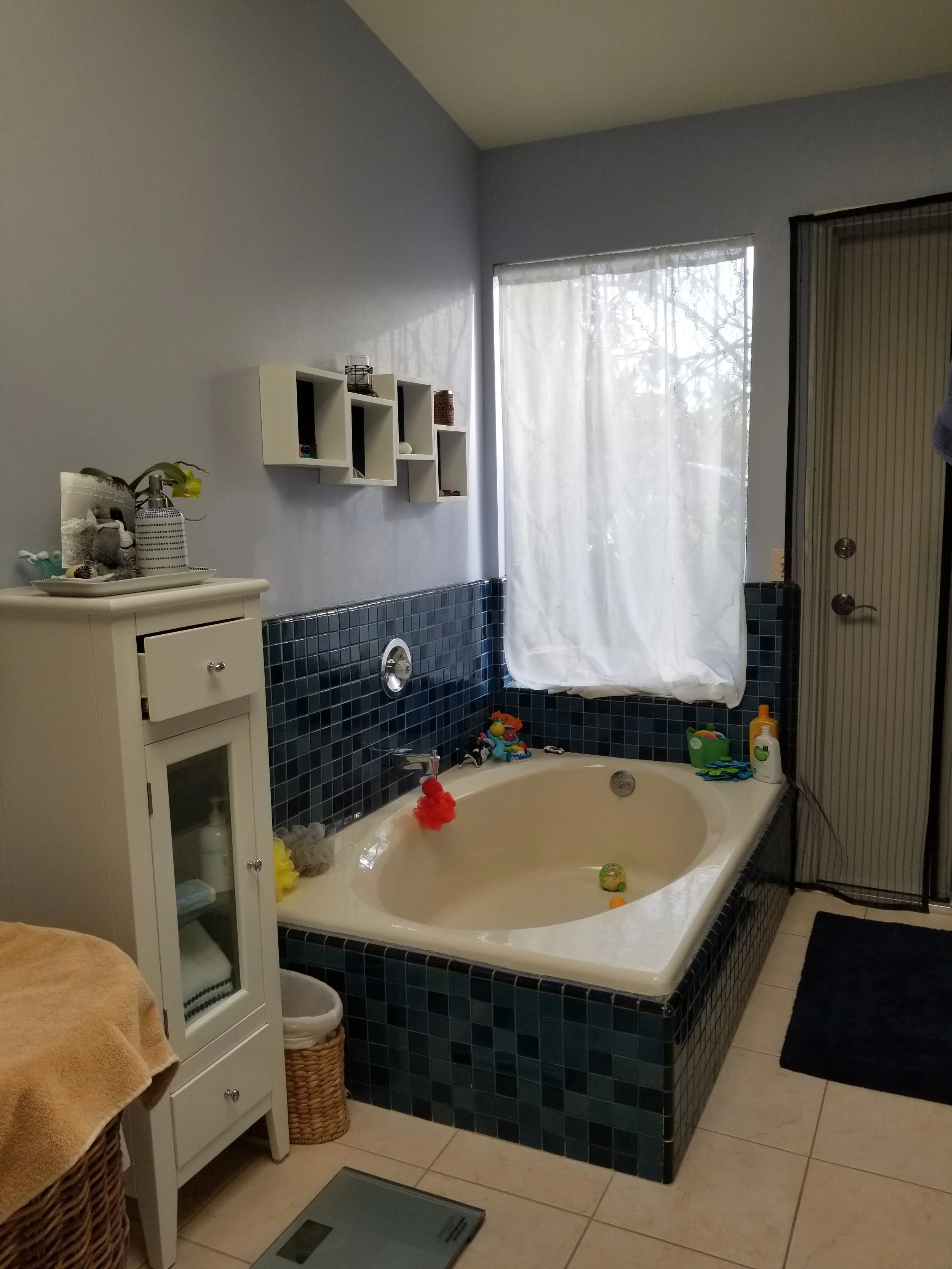 A bathroom with a bathtub surrounded by blue tiles, a window with white sheer curtains, a white cabinet with a mirror, and various children's toys on the edge of the tub.