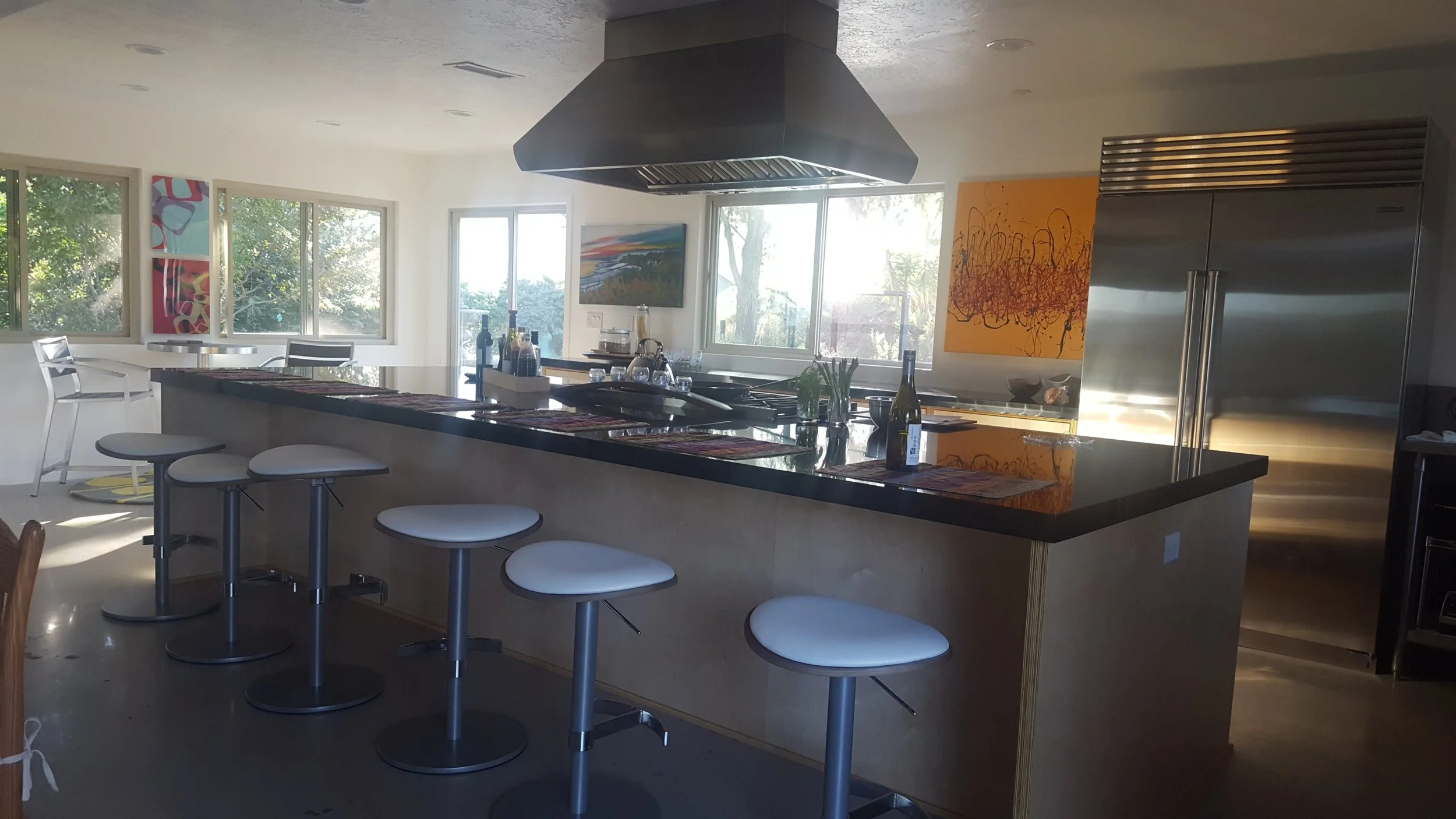 Bright kitchen with large central island, white stools, artistic paintings on walls, and large windows providing natural light.