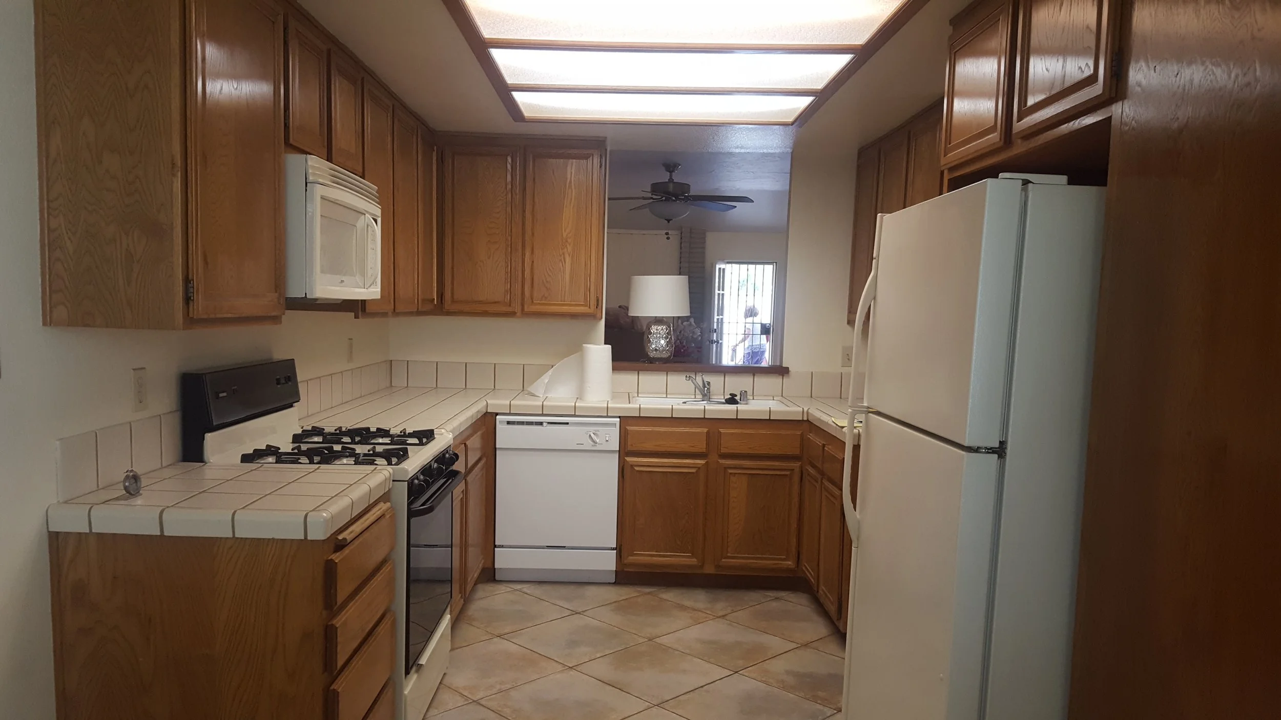 Kitchen with wooden cabinets, white appliances, beige tiled countertops and floor, fluorescent ceiling lights, window with a screen, ceiling fan in the adjacent room.