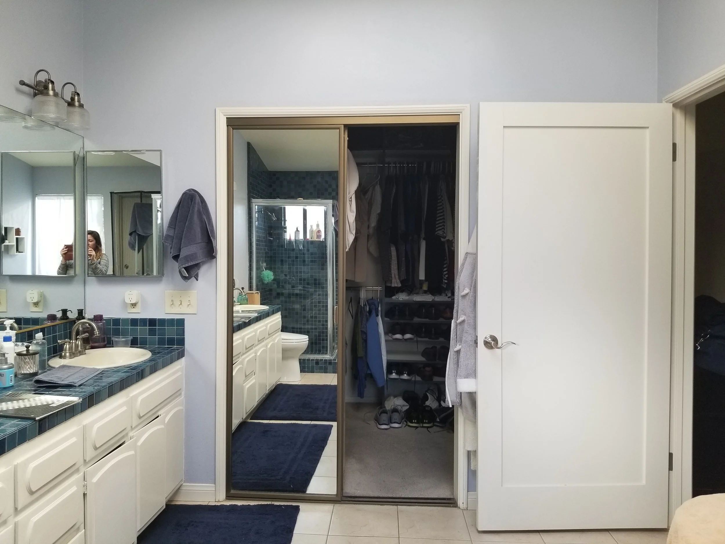 A bathroom with a white vanity, blue tile countertop, and a mirror. A towel hangs on a hook, and a woman is taking a photo in the mirror. There is a separate shower and closet visible, with clothes inside.