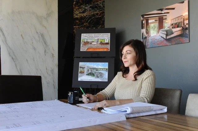 A woman sitting at a desk writing on large blueprints with two framed digital screens displaying construction site images behind her. There is wall art of a living room on the gray wall.