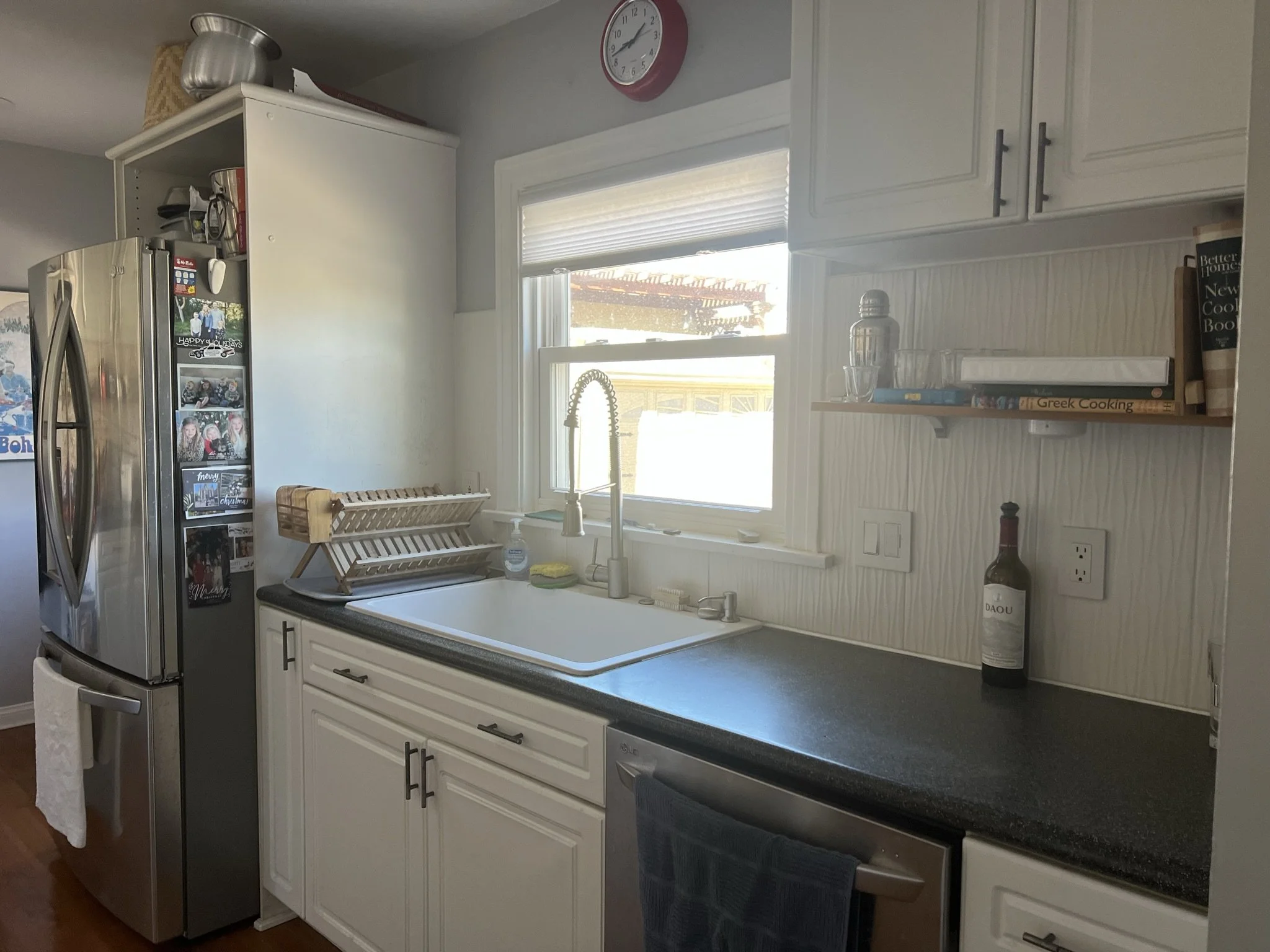 A kitchen with a stainless steel refrigerator, white cabinets, a window above the sink, a dish drying rack, a clock on the wall, and various items on the countertop and shelf.