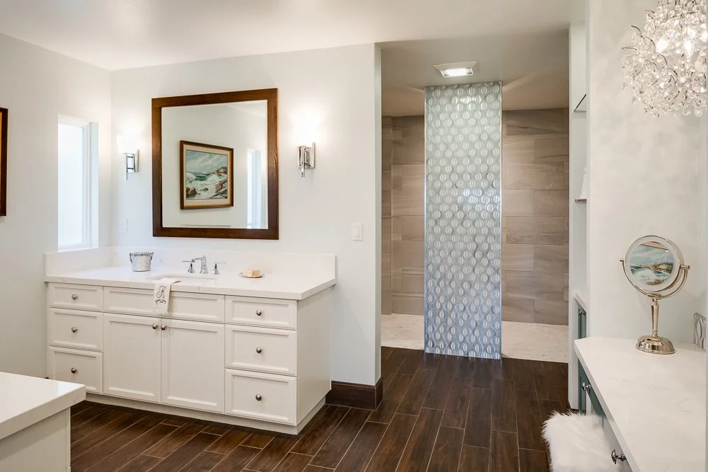 Bathroom with white vanity, large mirror, and wall-mounted lights, hardwood floor, walk-in shower with decorative partition, and a white and teal side table with mirror, decorative items, and fluffy stool.