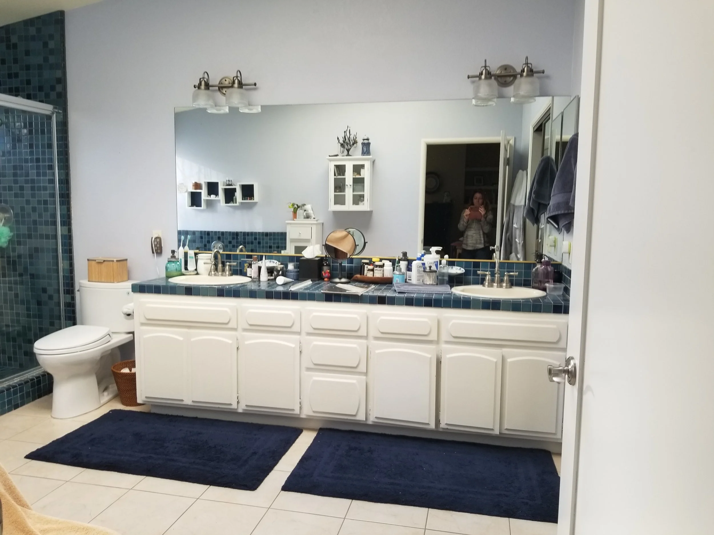 A bathroom with a large mirror above a double sink vanity, blue tile accents, a glass shower enclosure on the left, a toilet on the left, a dark blue rug on the tile floor, and a reflection of a woman taking the photo in the mirror.