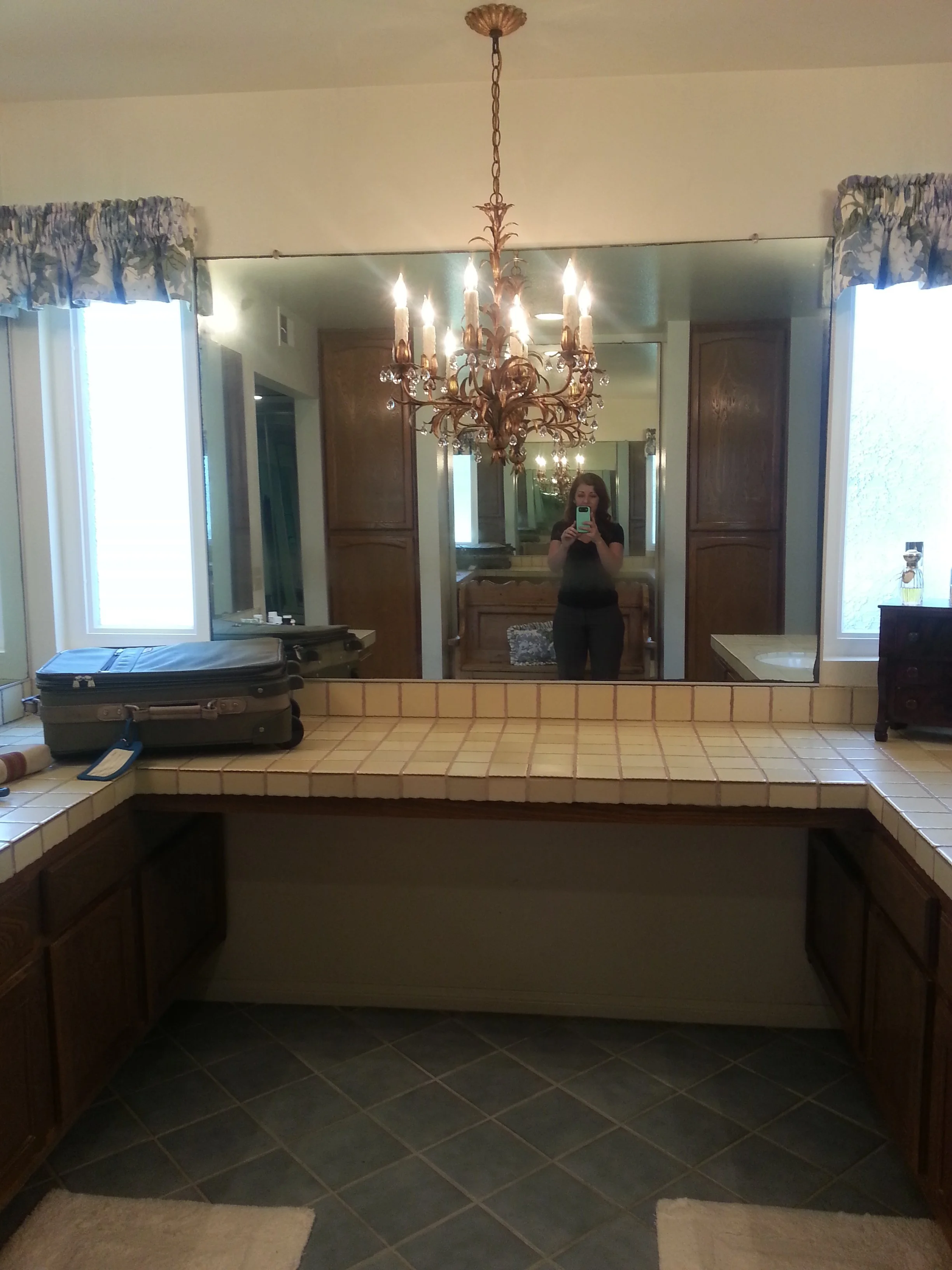 A bathroom vanity with a large mirror, a chandelier hanging from the ceiling, and a woman taking a selfie with her phone in the mirror. The counter has a suitcase and other items. There are two windows with floral valances, and dark tiled flooring.