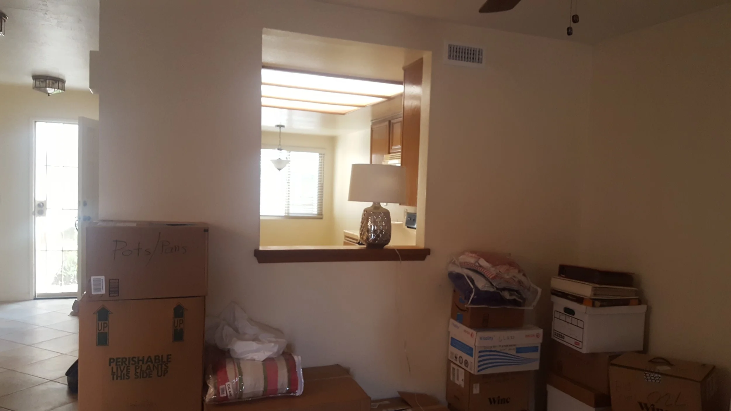 Living room with moving boxes and packing supplies, including a large box labeled for fragile live plants, a blanket rolled on the floor, and a small table with a lamp. The room has a view through an open door and a window with blinds.