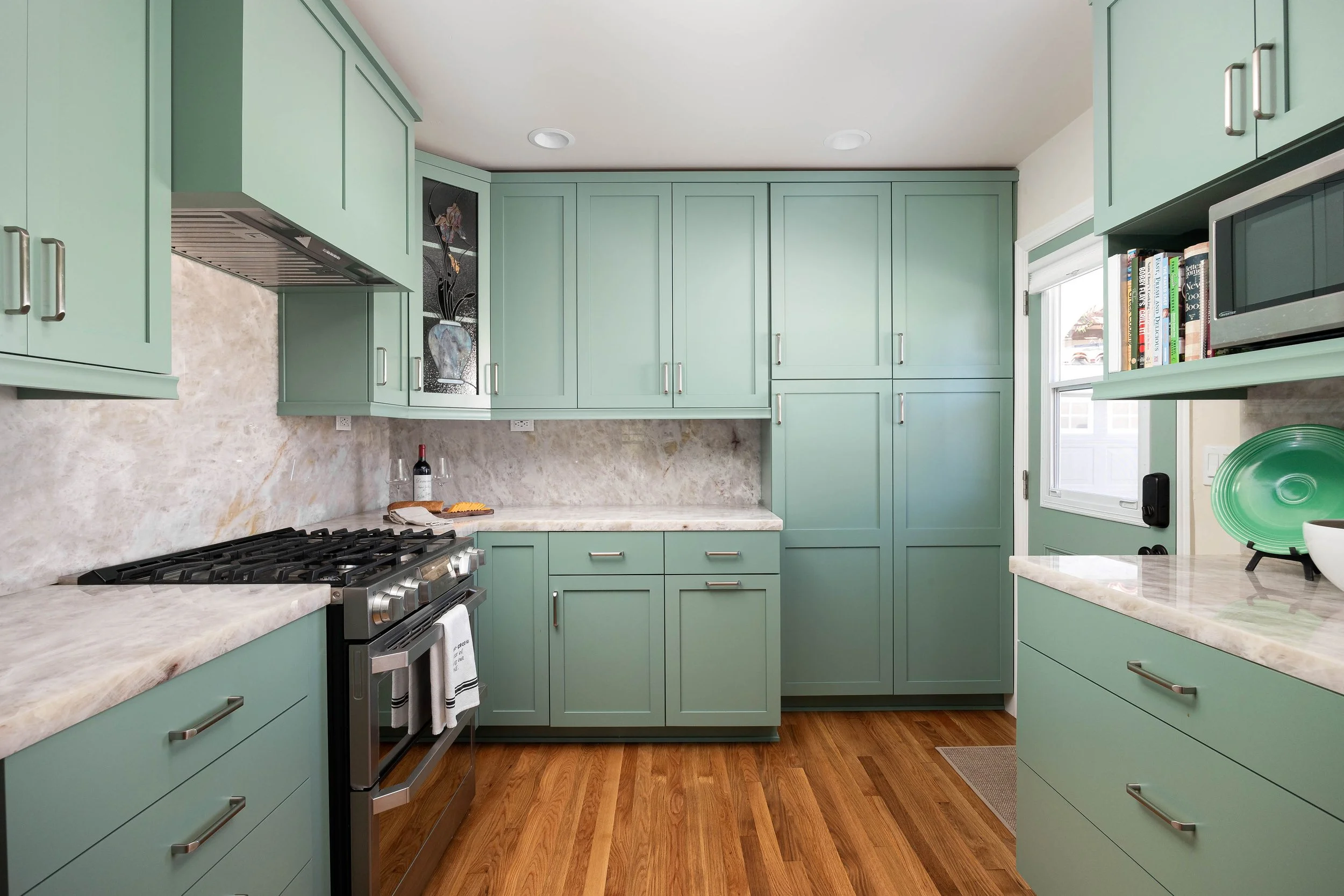 A kitchen with green cabinets, a marble countertop, a gas stove, a window, books on a shelf, and hardwood floors.