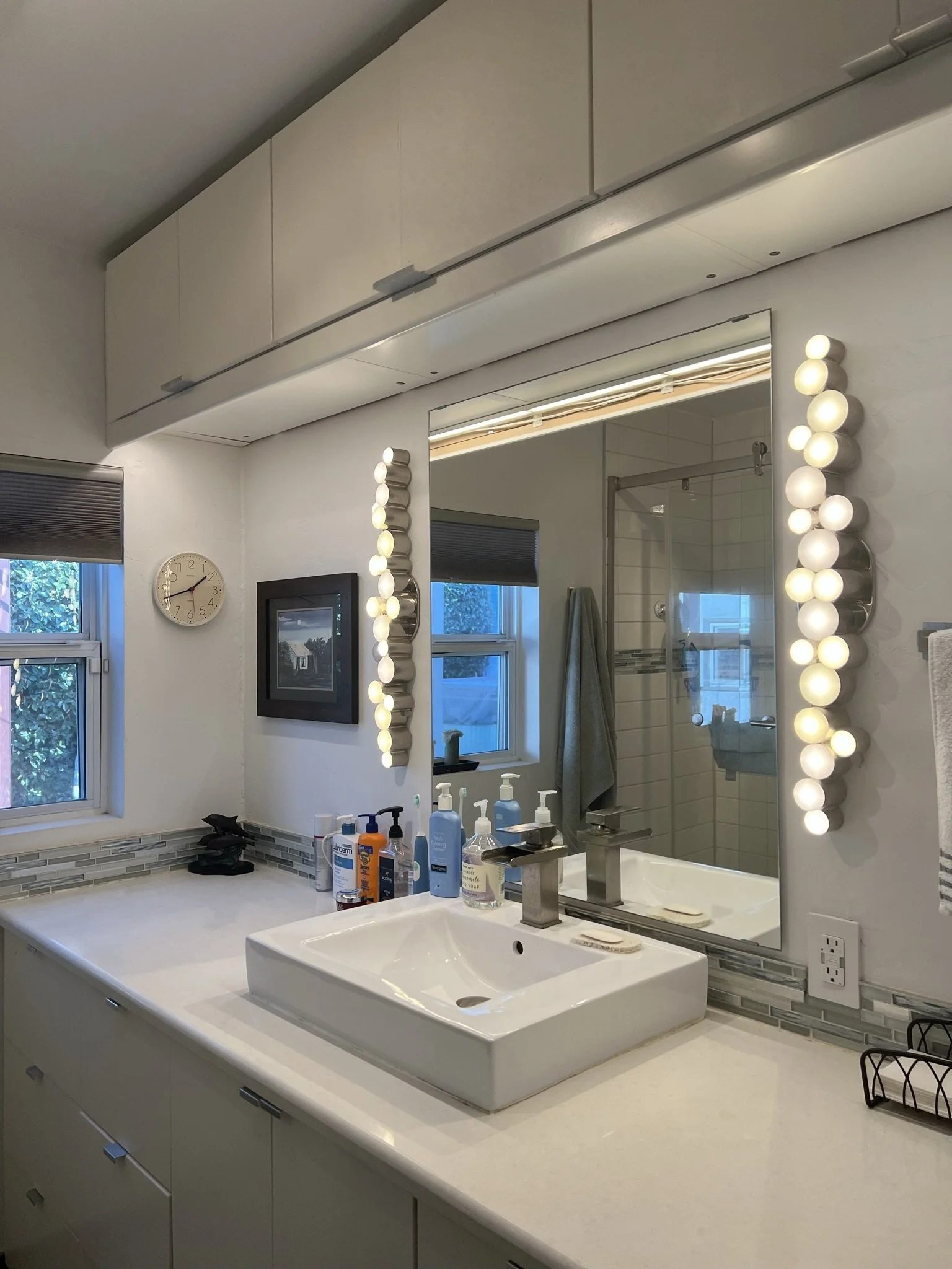 Modern bathroom with a white countertop, rectangular sink, and various soap bottles. Large mirror with decorative light bulbs on either side, and a window with a dark blind.