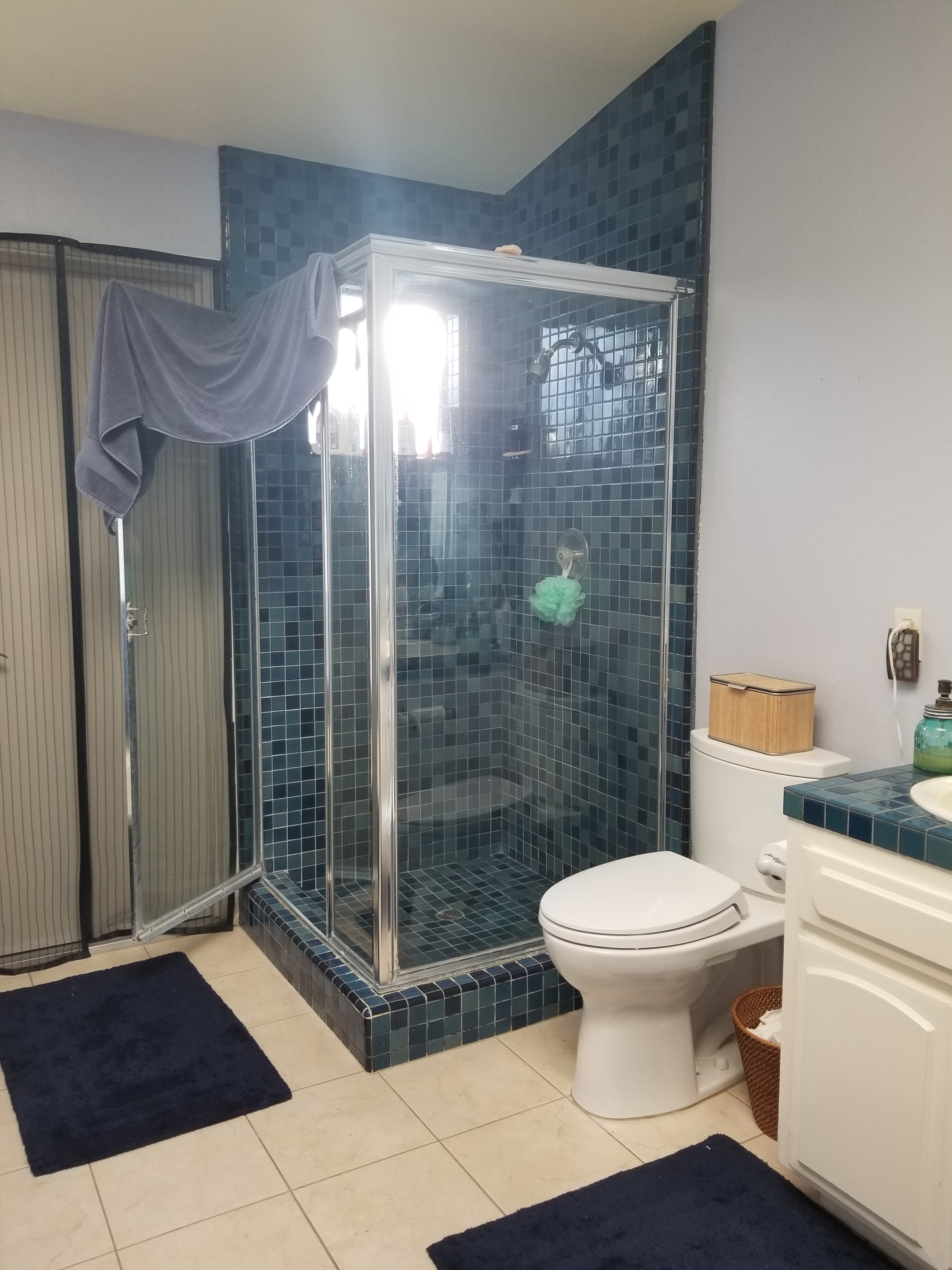 A bathroom with a glass shower enclosure with blue tiles, a toilet, a white vanity, and navy blue rugs on beige tiled floor.
