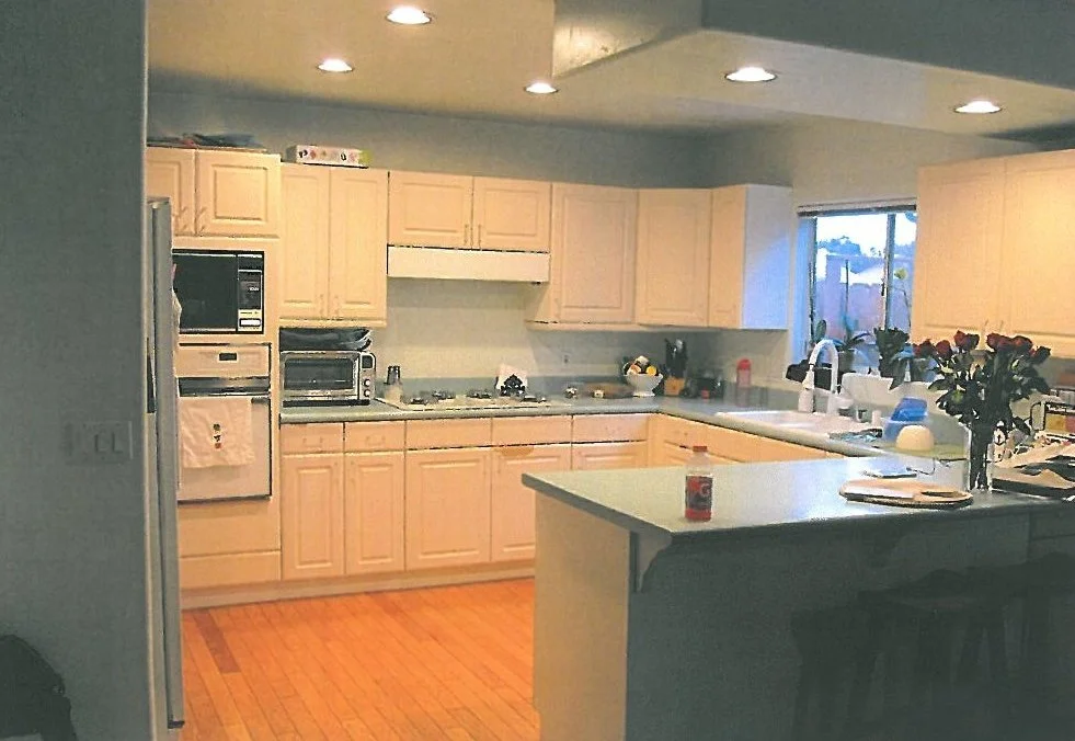 Kitchen with beige cabinets, white countertops, and wooden flooring. Several appliances, a double sink, and a window with potted plants are visible. Items like flowers, bottles, and papers are on the counter.