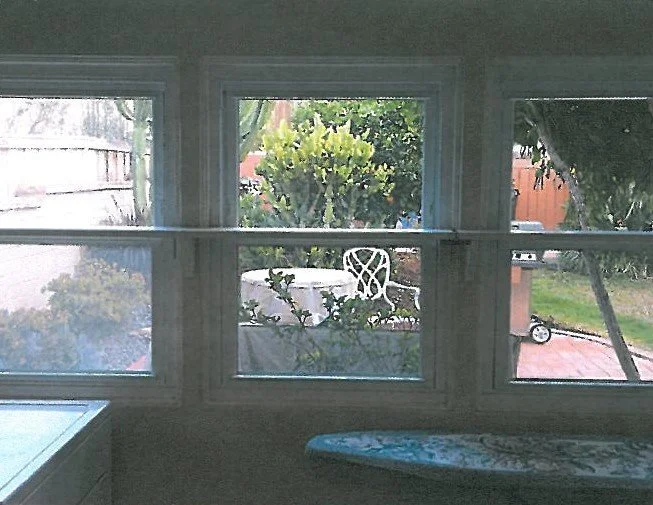 View through a three-pane window showing a backyard with bushes, trees, a patio, a small white table, and a white chair outside.