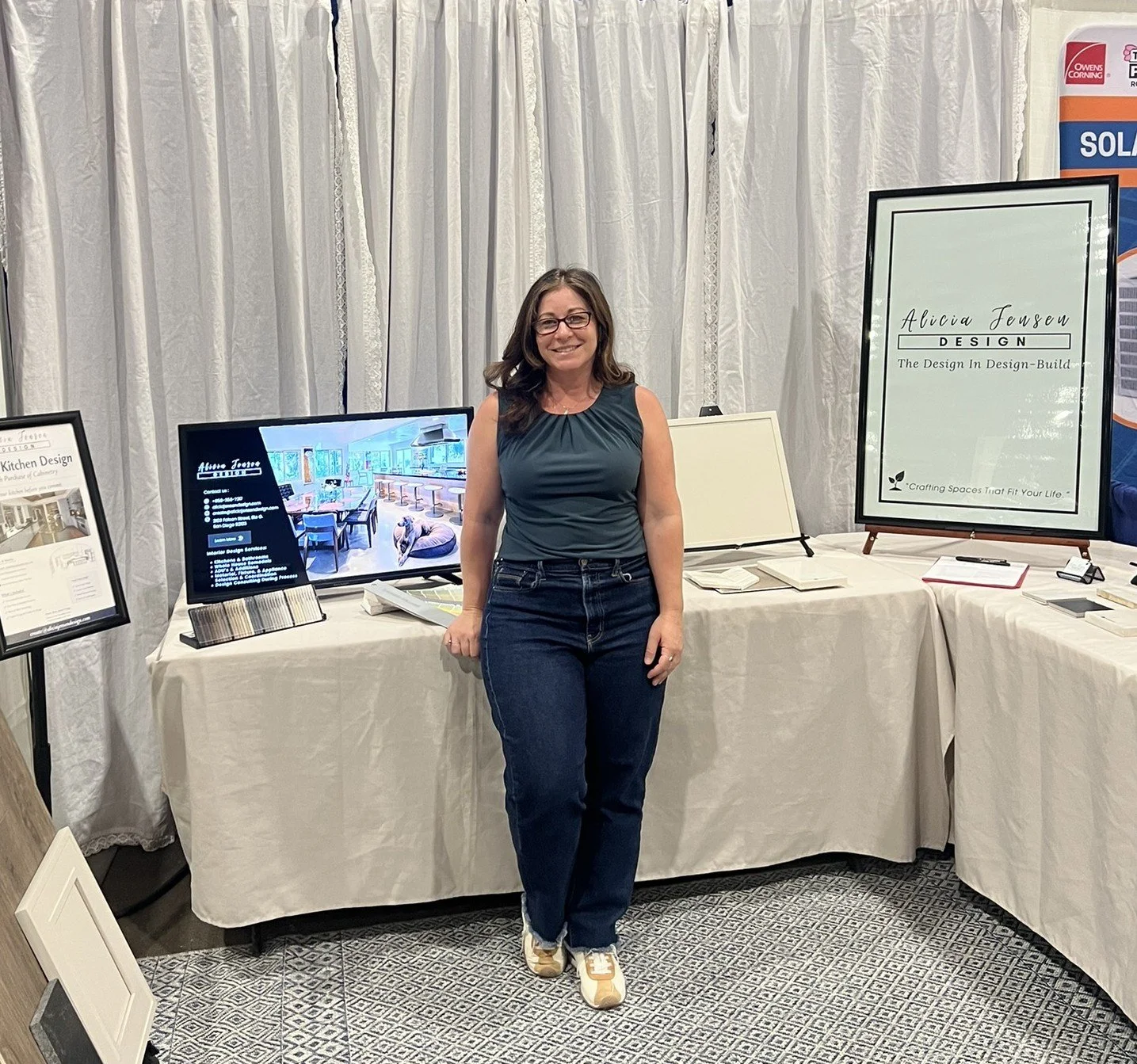 Woman standing in front of a display booth at an event, with a large framed poster that reads 'Alicia Jensen Design' and shows a tagline about design and build, surrounded by posters and product samples.