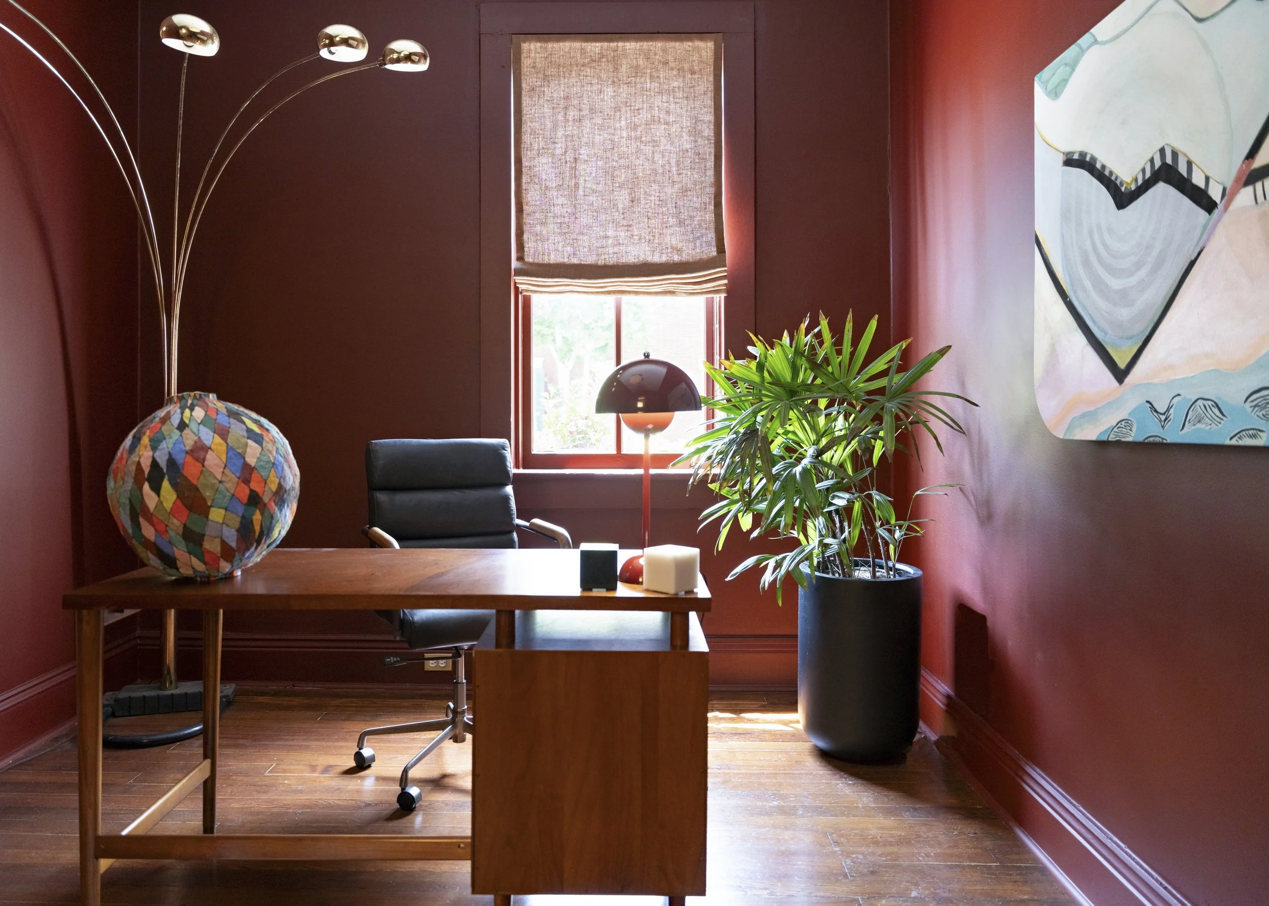 A cozy living room with red painted walls and wooden floors. There is a wooden dining table with a black office chair, a colorful patterned lamp, a modern black desk lamp, a large potted plant, and abstract wall art. A window with a woven shade lets in natural light.