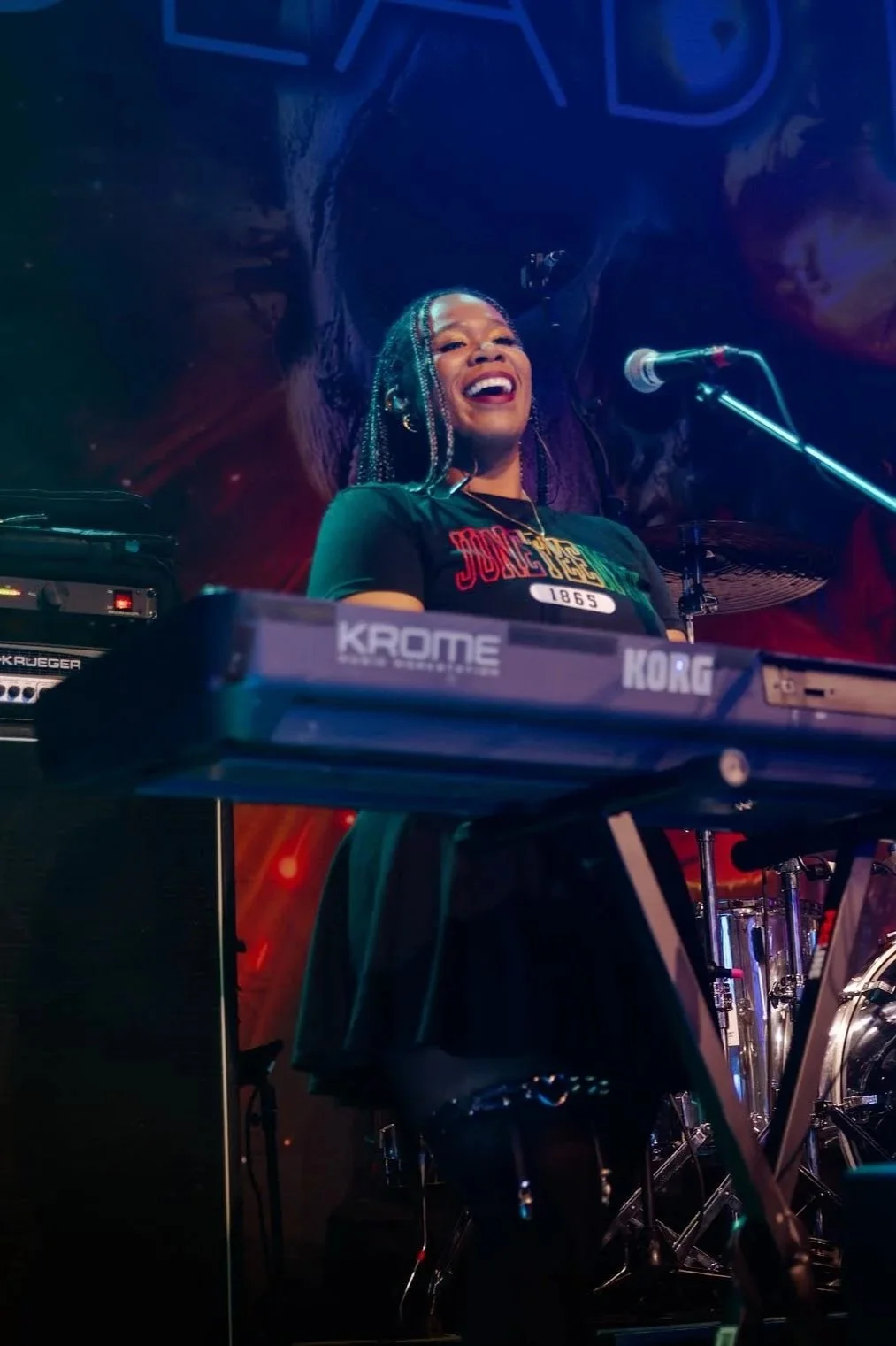 A woman with braided hair and a bright smile playing a Korg keyboard on stage, with a microphone stand nearby, and a dark, colorful background.