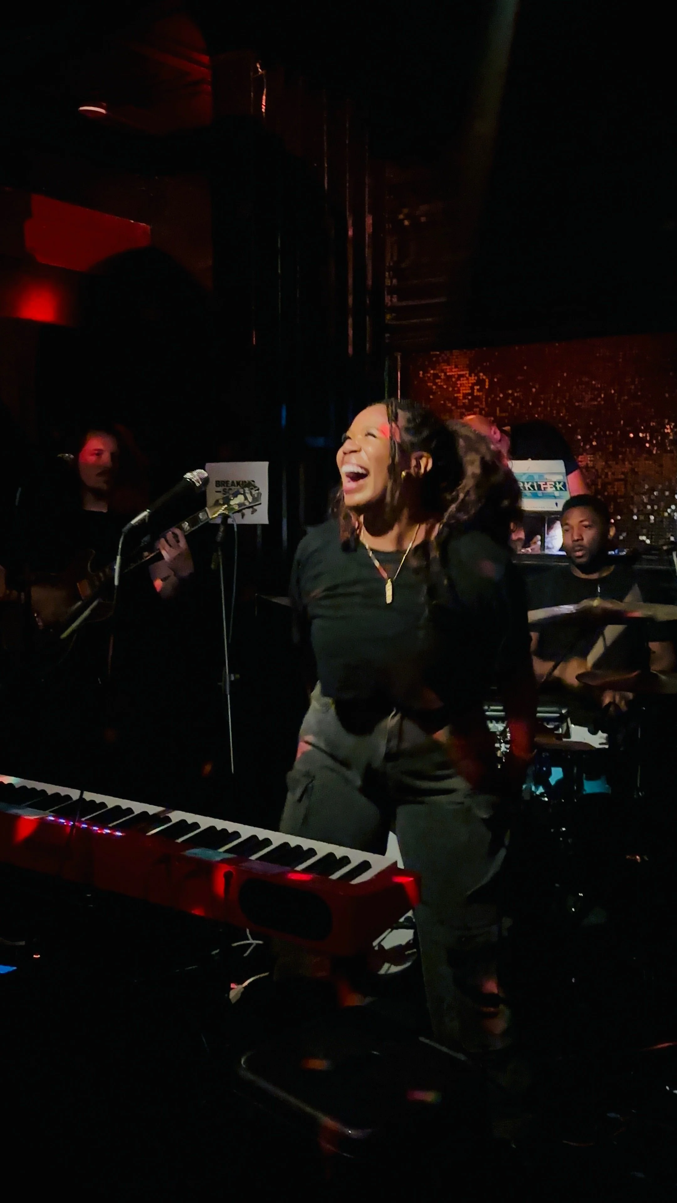 A woman singing and dancing in a dimly lit music venue, with a pianist keyboard in the foreground, a guitarist, and a drummer in the background.
