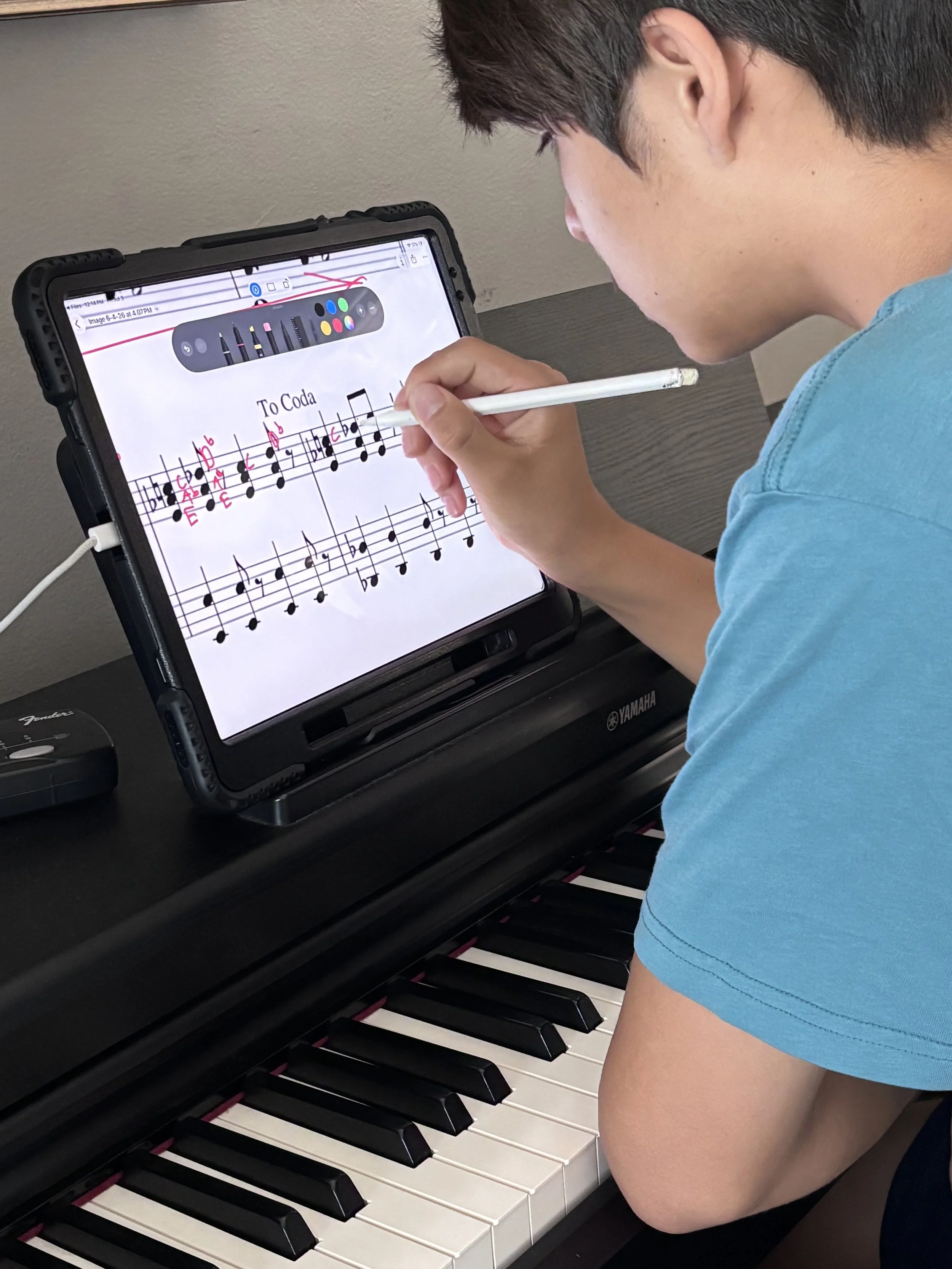 A young boy is playing a digital piano and using a tablet to view sheet music. The tablet displays a music score with a stylus in his right hand.