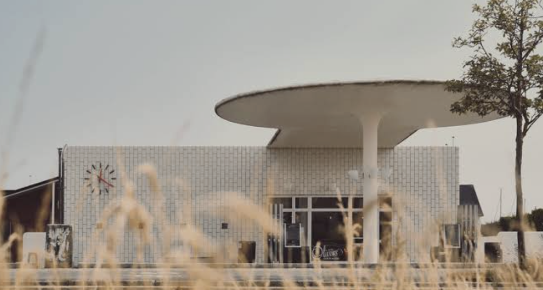 Modern building with a distinctive round, overhanging roof, a large clock on the exterior wall, and a grassy foreground with tall dry grass and a tree on the right side.