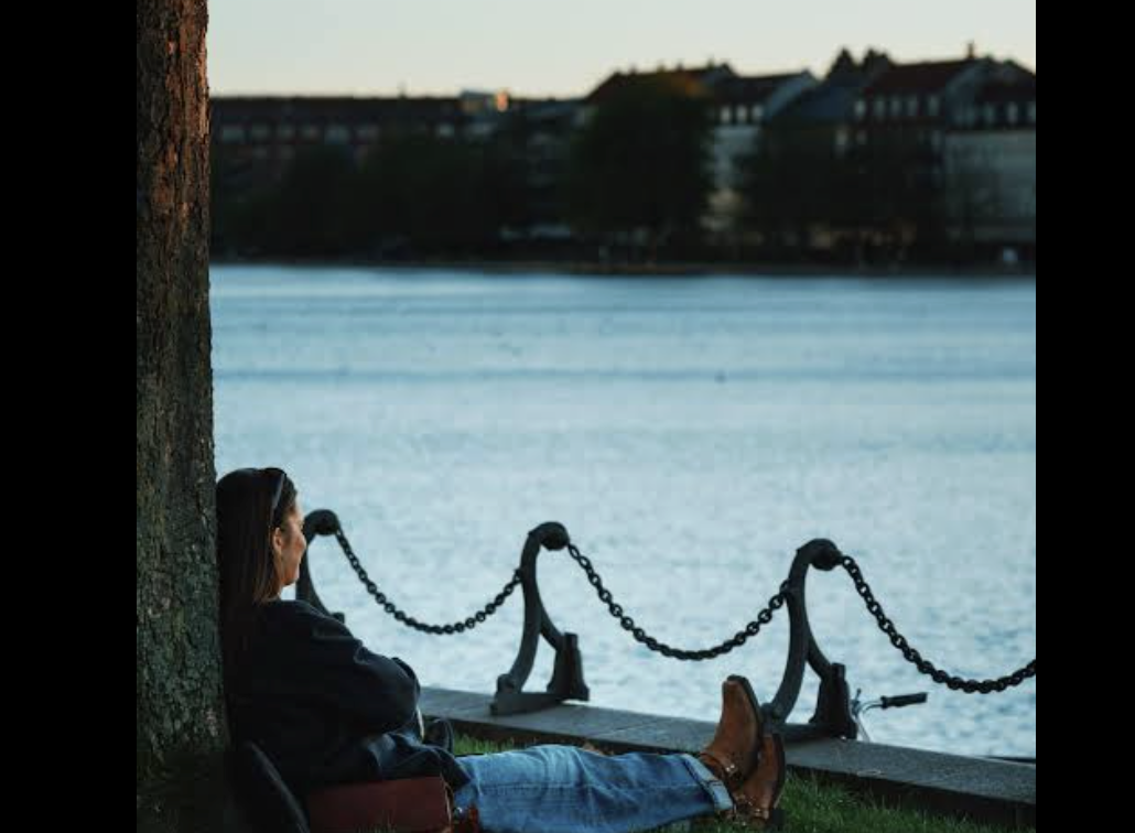 A woman sitting on the grass leaning against a tree, looking across a body of water during sunset with buildings in the background.