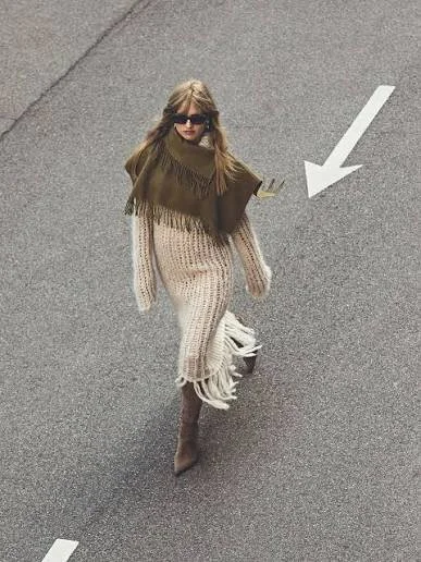 A woman walking on a gray road, wearing sunglasses, a brown fringed shawl, a beige knit dress, and brown boots.
