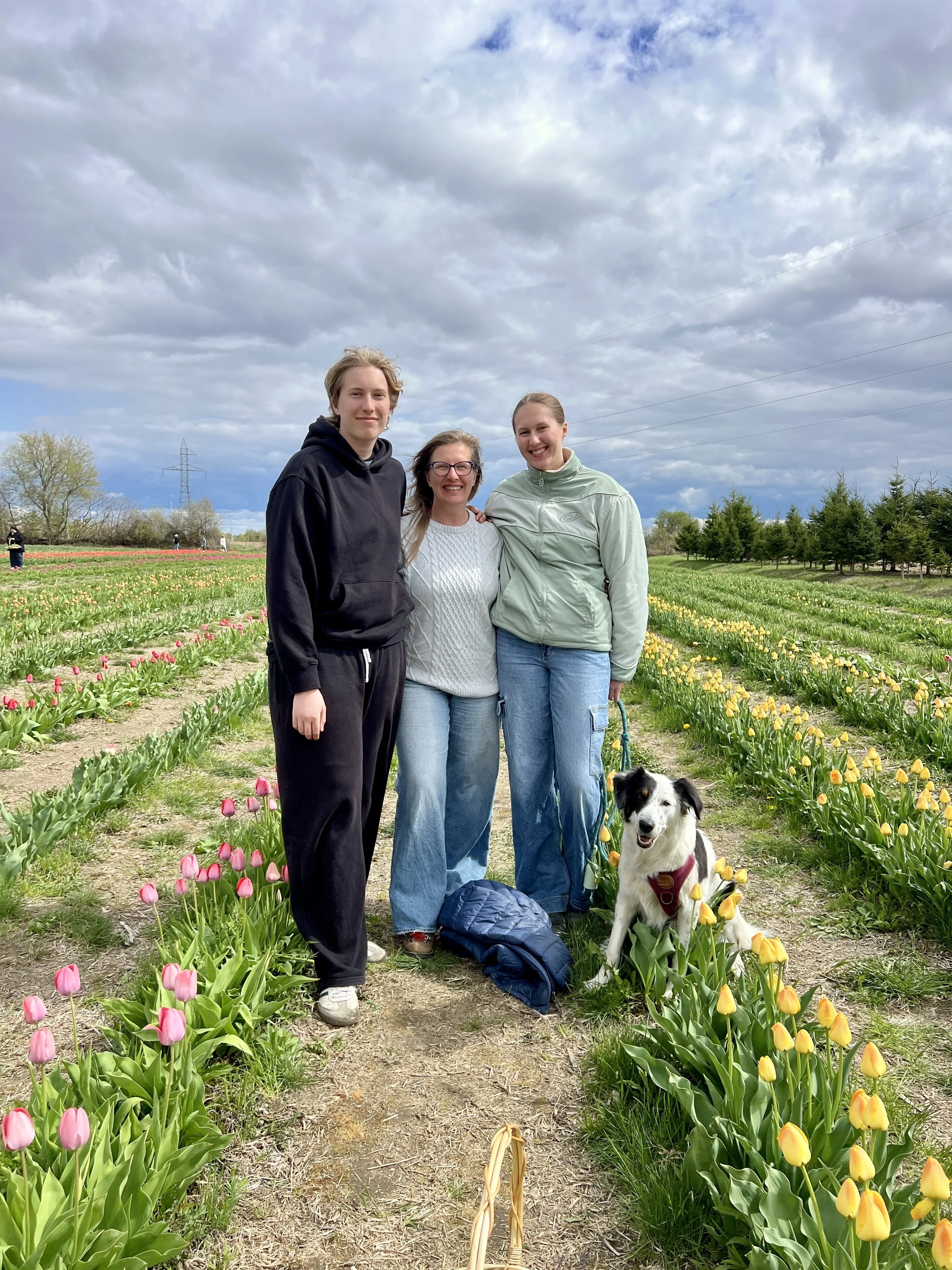 Tatiana Doula with her 2 teens and a dog standing on a dirt path in a tulip field with colorful tulips, trees, power lines, and a cloudy sky in the background.