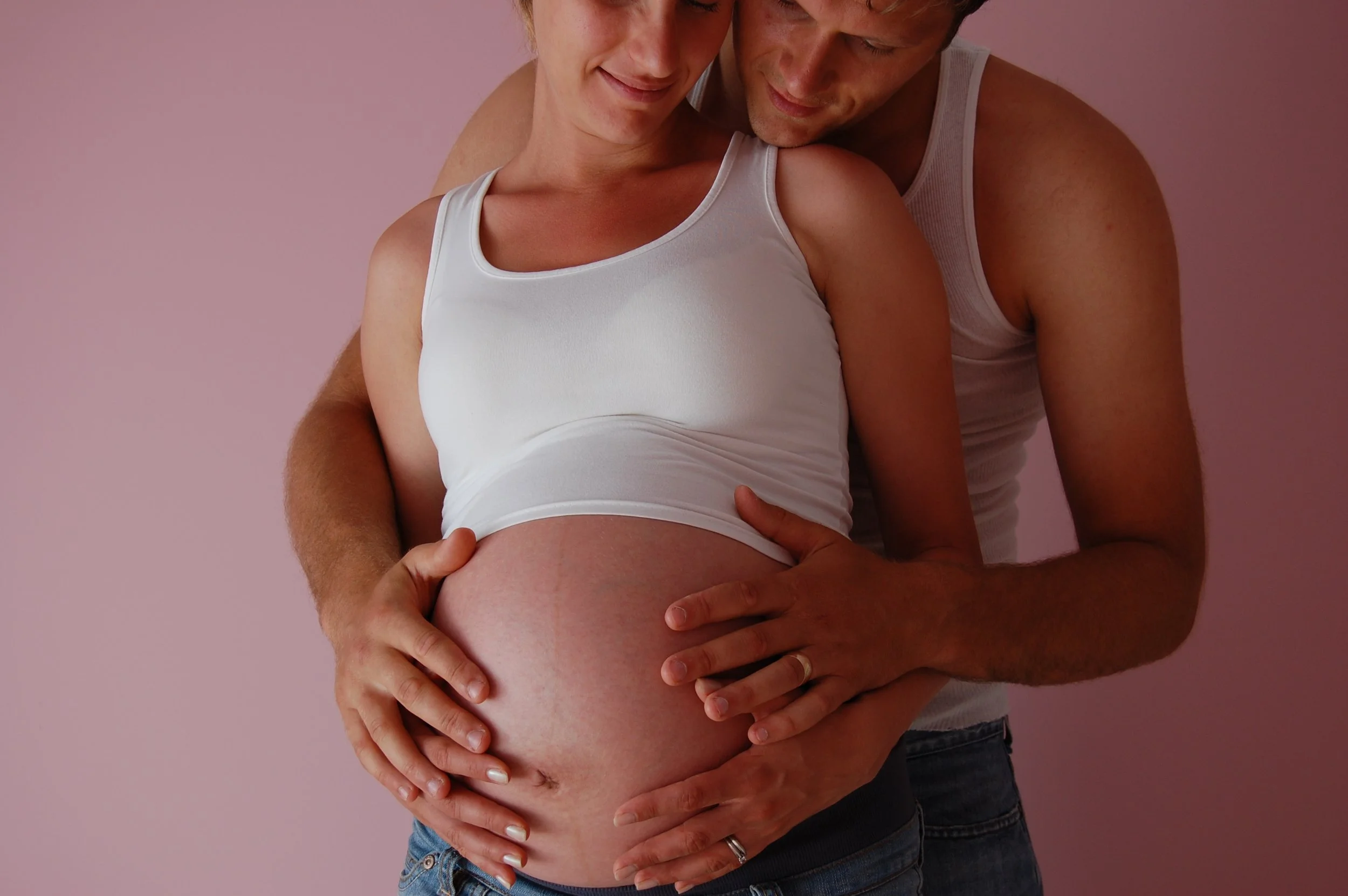 A pregnant woman in a white tank top and a man in a white sleeveless shirt hugging and holding her belly against a light pink background.