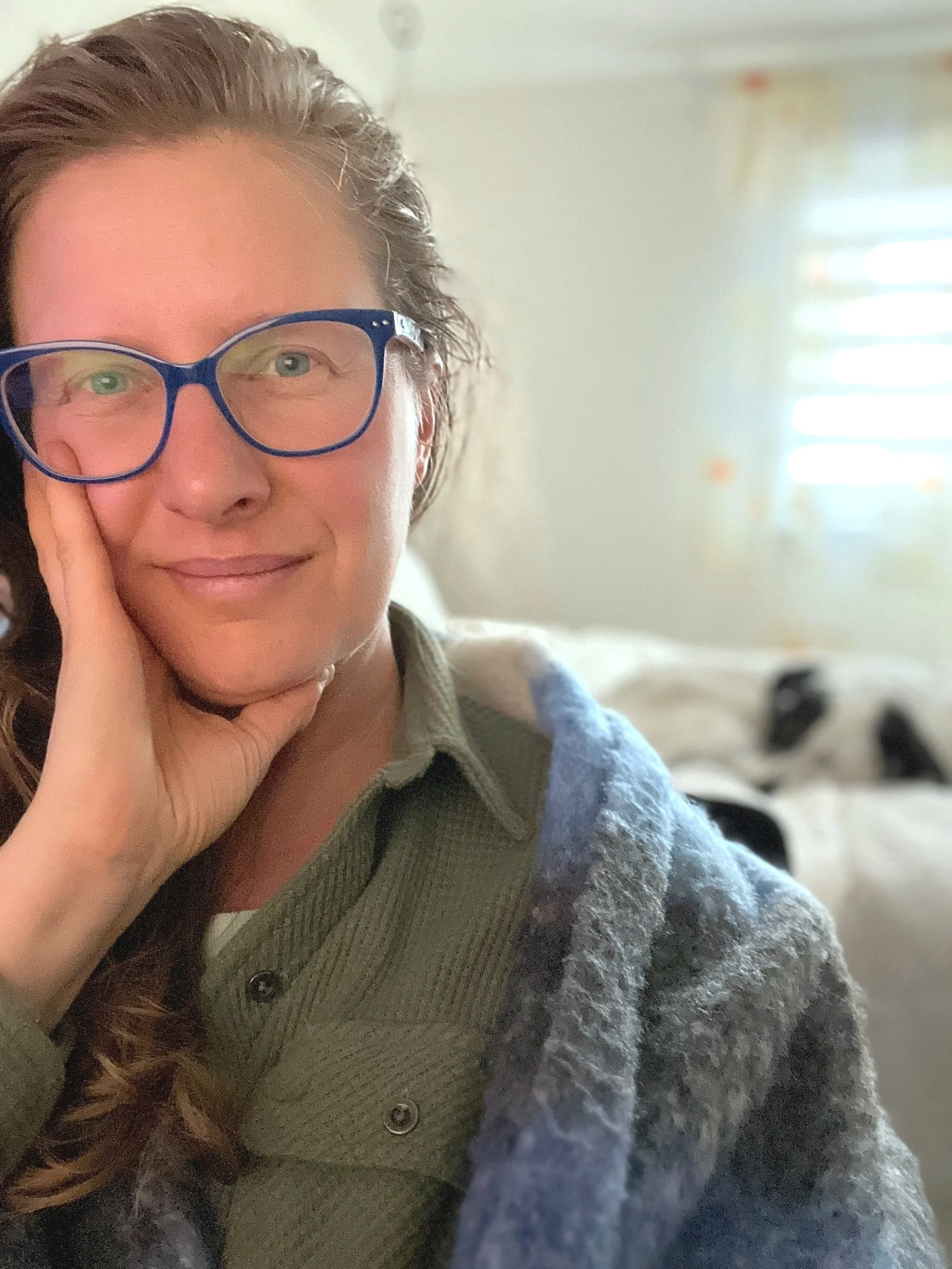 Close-up of a welcoming doula wearing blue glasses, smiling with her hand resting on her face, in a room with sunlight coming through blinds.
