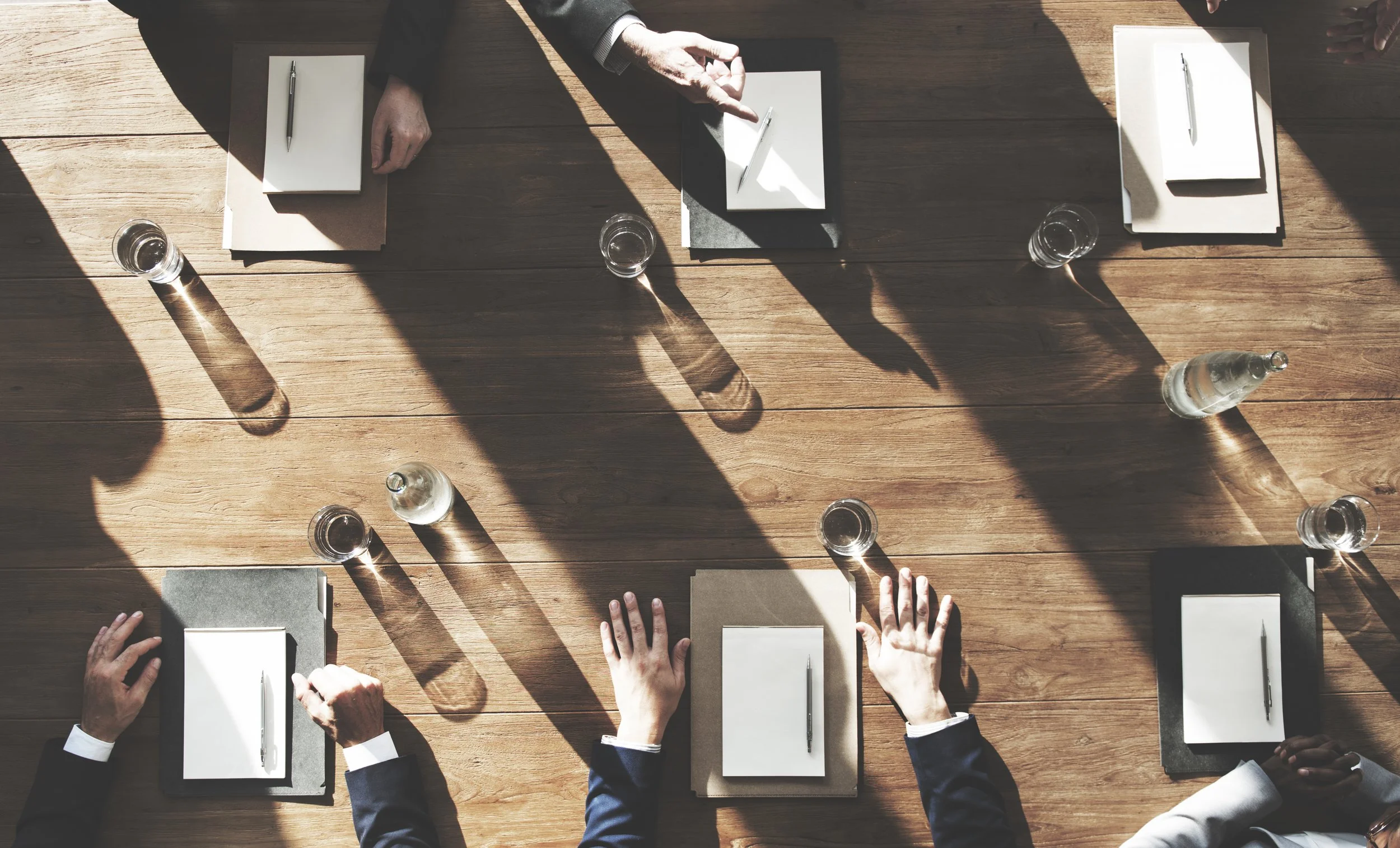 A top-down view of a business meeting with several people wearing business suits seated around a wooden table. There are notebooks, pens, and glasses of water on the table. Shadows from sunlight are cast across the table.