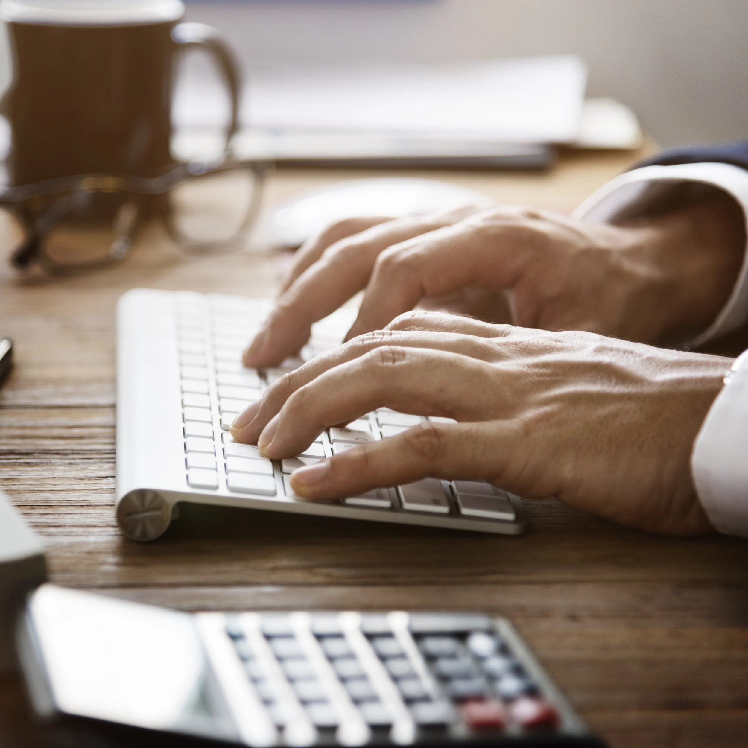 Close-up of hands typing on a keyboard on a wooden desk with a blurred background of a brown mug, papers, and a laptop.
