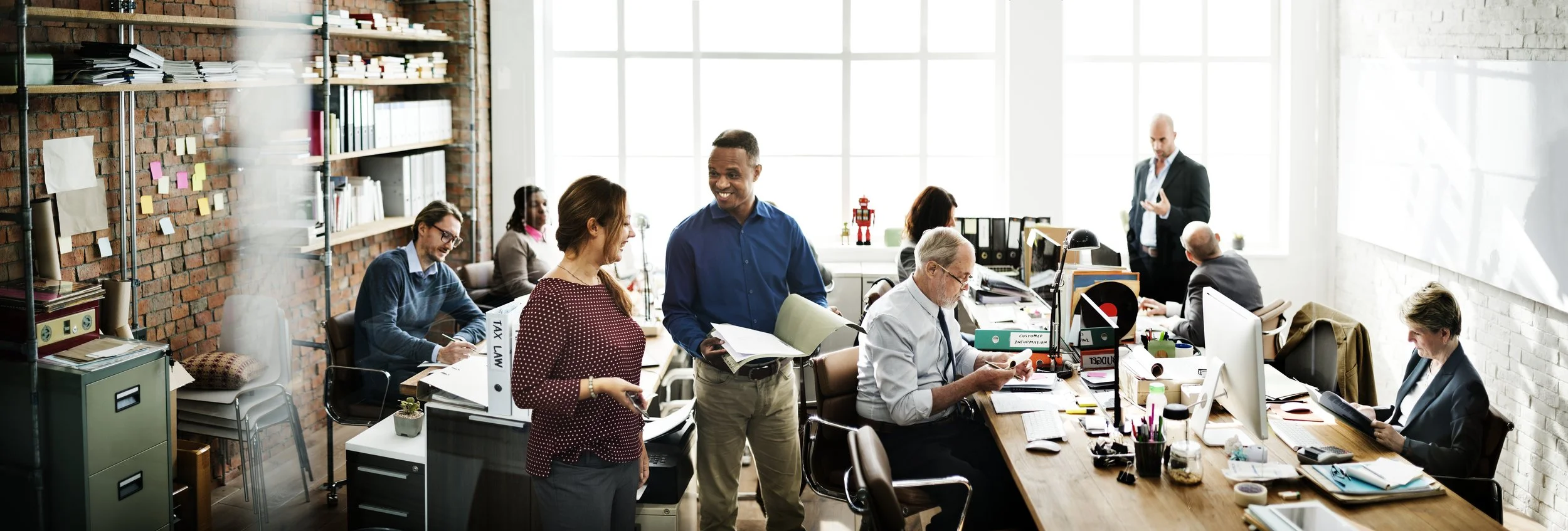 Office scene with diverse team members working and chatting, with large windows and exposed brick walls.