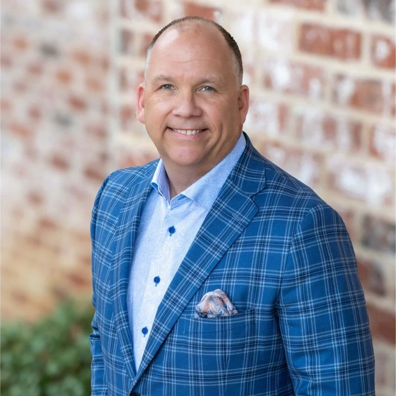 A smiling man in a blue plaid suit with a light blue shirt standing outdoors against a brick wall.