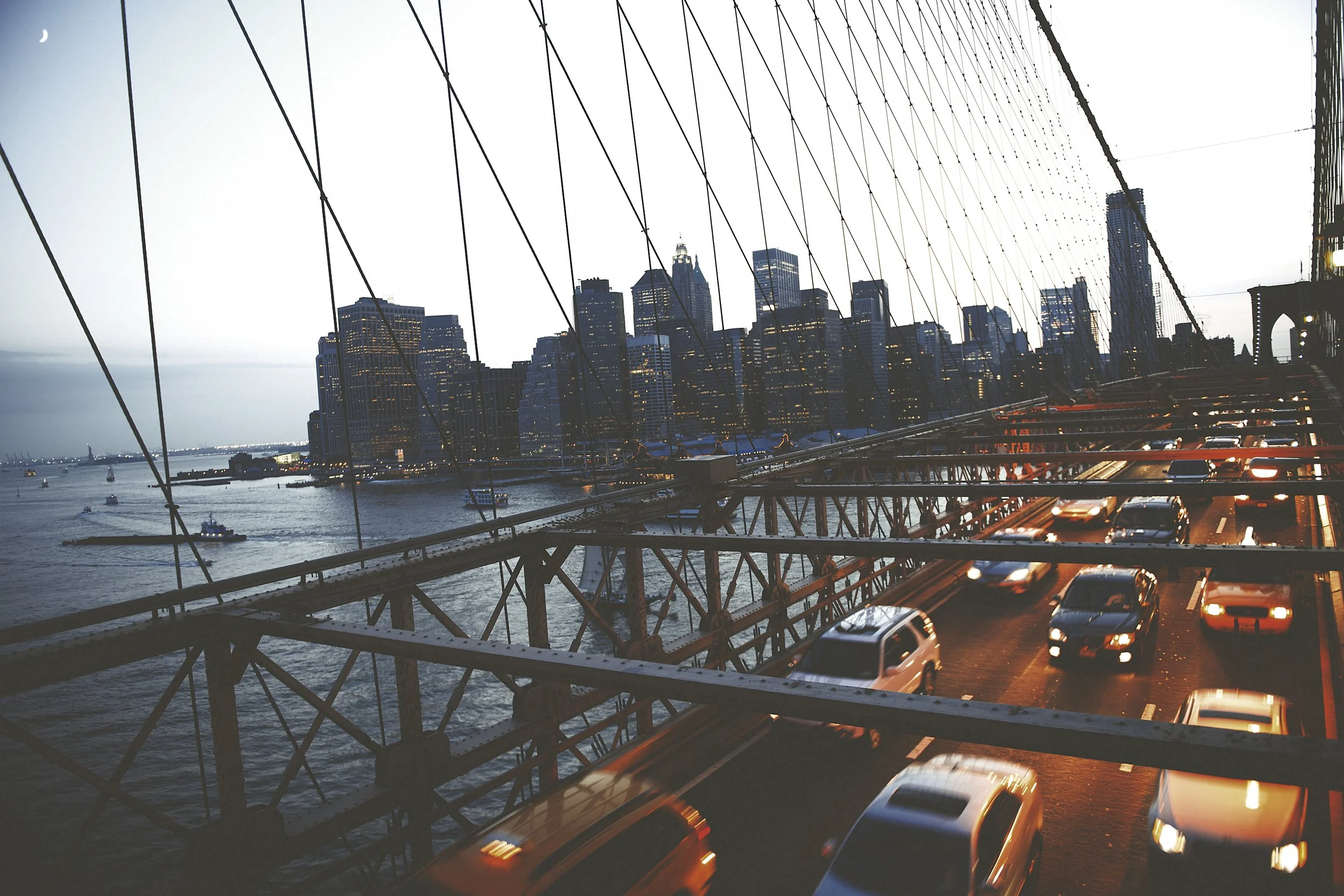 View of the Brooklyn Bridge at dusk with the Manhattan skyline and the Statue of Liberty across the water, cars driving on the bridge, and a crescent moon in the sky.