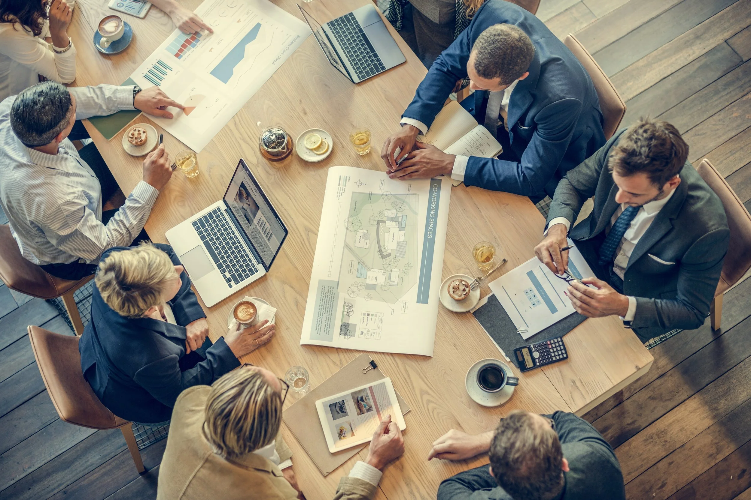 Business meeting with several individuals around a wooden table discussing charts, graphs, and architectural plans. Drinks and snacks are on the table, and laptops and tablets are open.